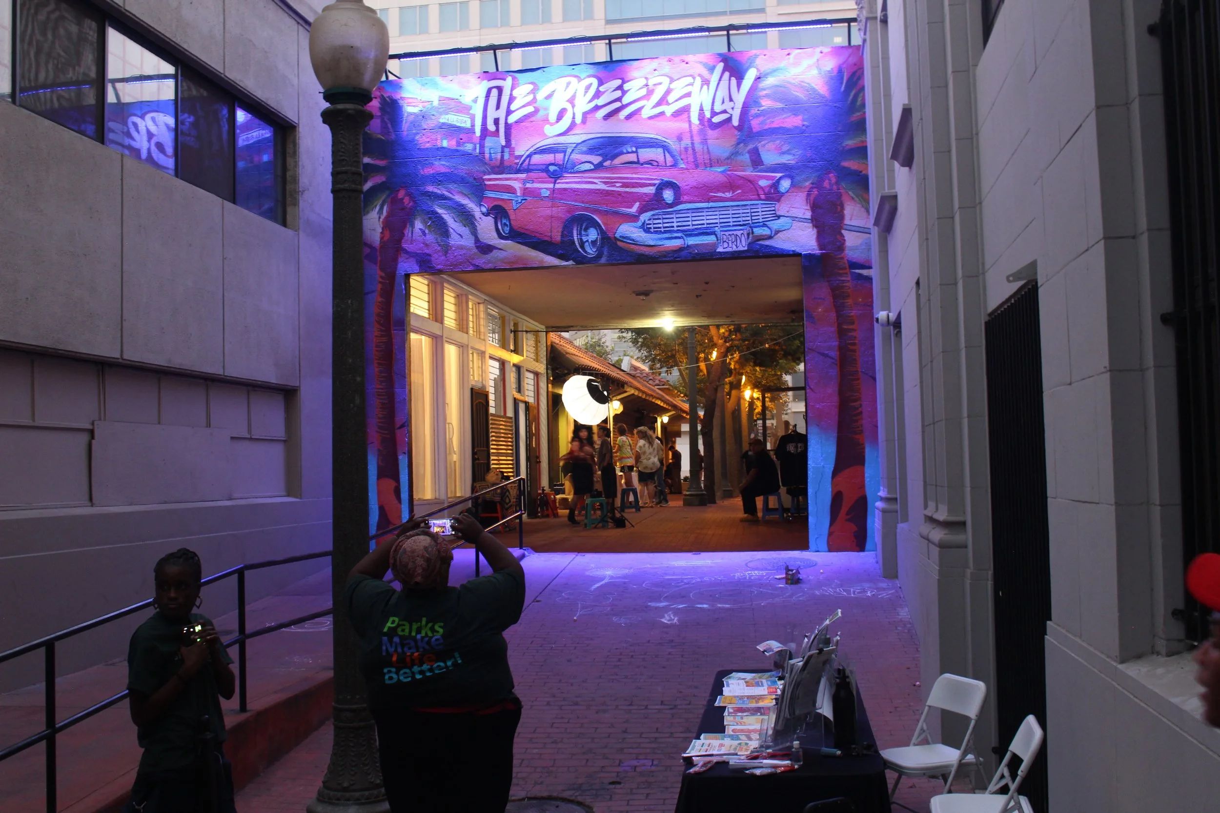 An outdoor scene at dusk with a colorful mural of a vintage car and palm trees, a group of people gathering under a canopy, and someone taking a photo in front. There are chairs and a table with flyers, illuminated by warm lights.