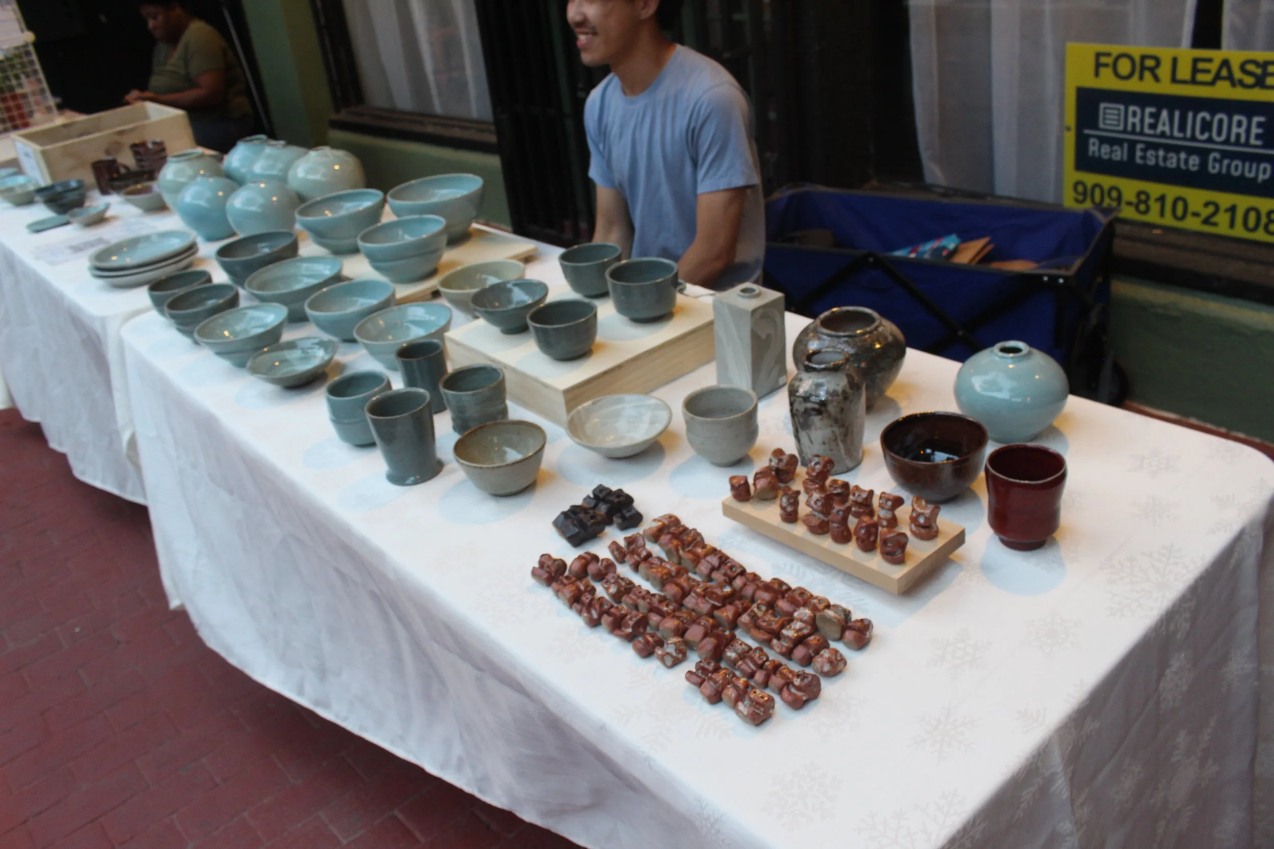 Table displaying various ceramic pottery items including bowls, vases, and cups at an indoor market, with a person behind the table and a yellow real estate for lease sign in the background.
