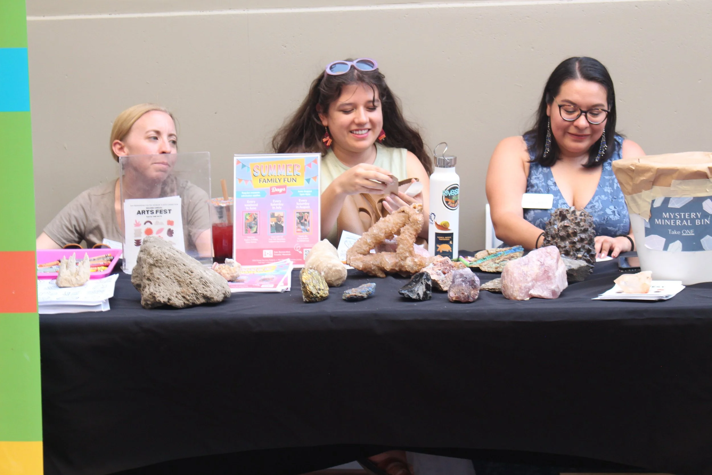 Three women sitting at a table with rocks and minerals on display, at a mineral and rock exhibit or event.