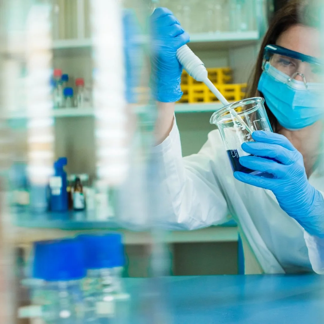 A scientist working in a laboratory, wearing gloves, a face mask, and protective glasses, using a pipette to transfer liquid into a test tube.