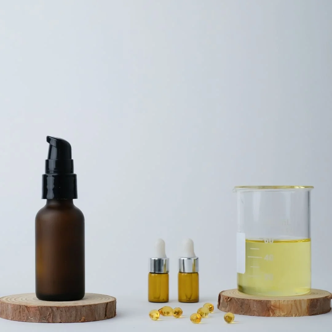 A collection of skincare products, including a dark brown pump bottle on the left, two small amber glass dropper bottles in the center, a glass beaker filled with yellow liquid on the right, and small yellow capsules scattered in front, all displayed on wooden coasters against a white background.