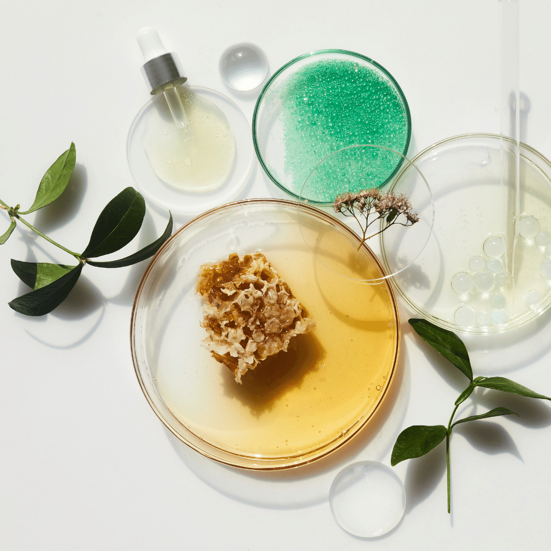 Assorted skincare products and natural ingredients on a white surface, including a dropper bottle, a green gel in a glass dish, clear capsules, a bar of soap or scrub, and green leaves.