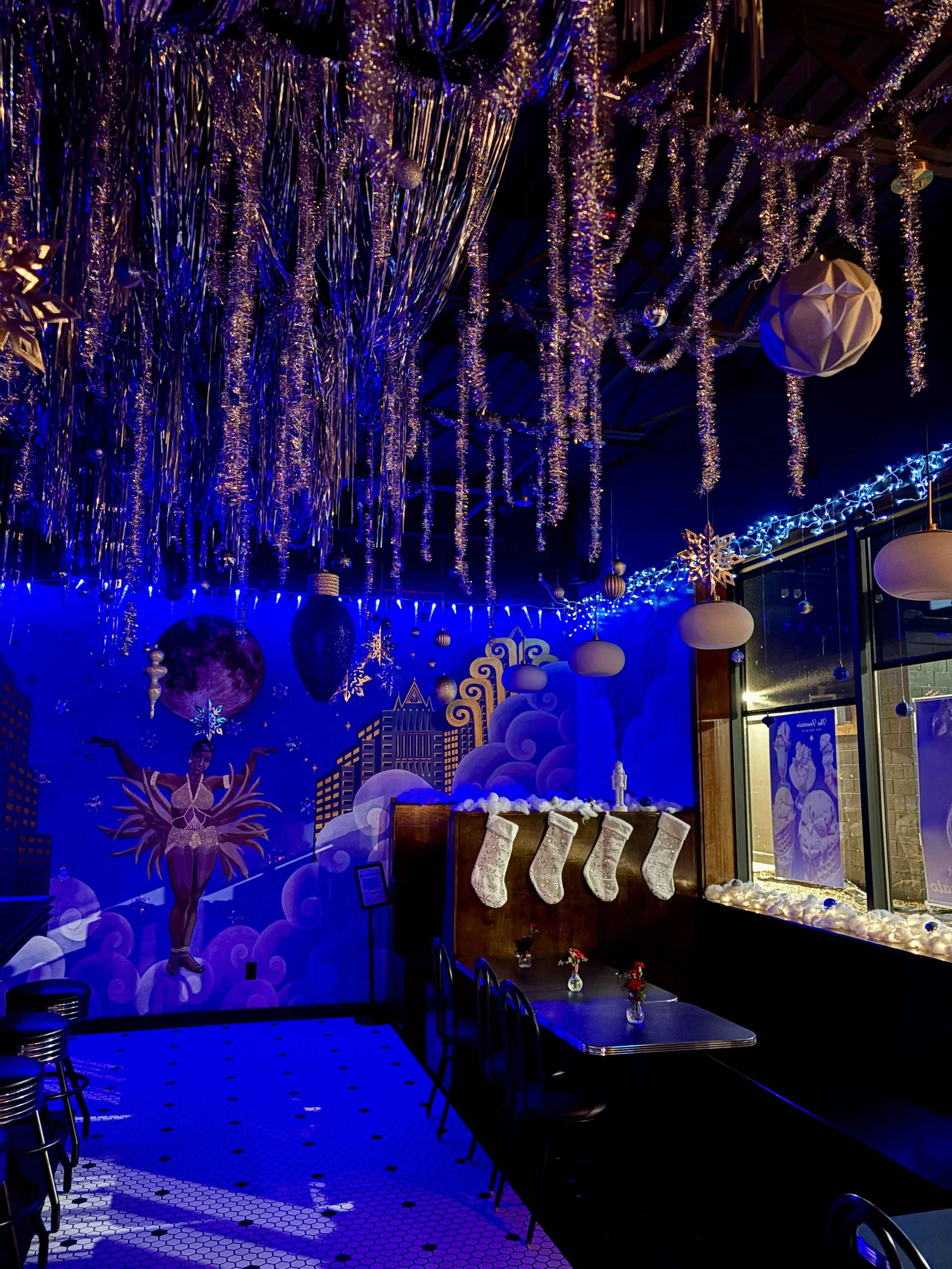 The Iced Fountain Pop Up: Winter-themed diner interior with blue lighting, tinsel decorations, stockings, and a mural of a dancer. Tables and stools present.