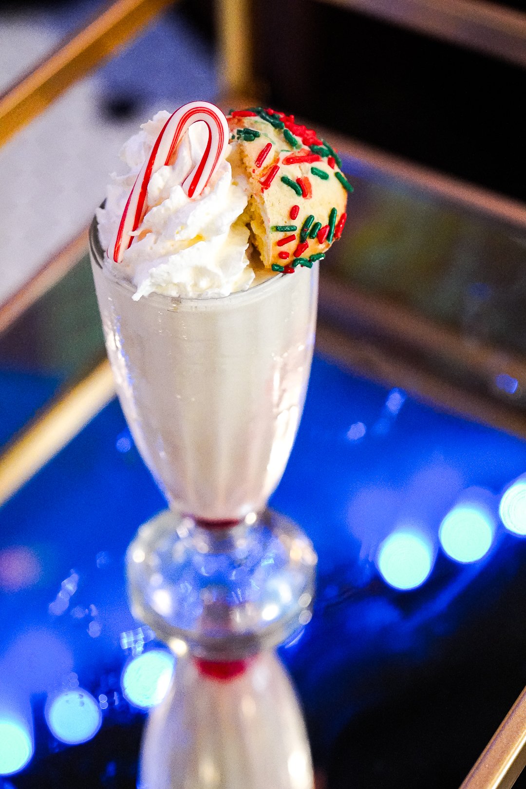 Gooey Butter Peppermint Milkshake in a glass with whipped cream, candy cane garnish, and a holiday cookie on top. Served on a reflective surface with blue lights.