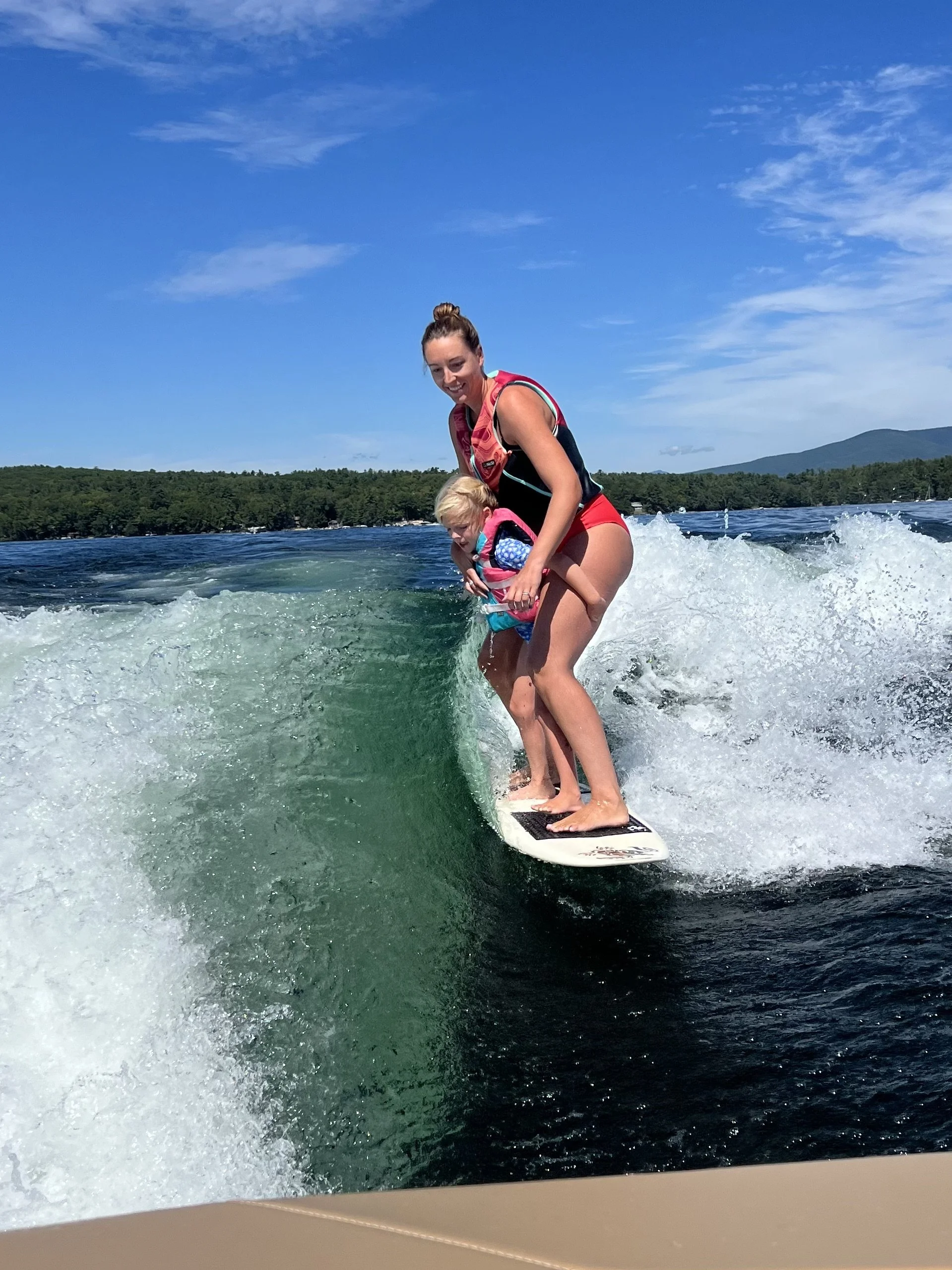 A woman and a young child riding a wakeboard on a lake during sunny weather with a clear blue sky and a forested shoreline in the background.