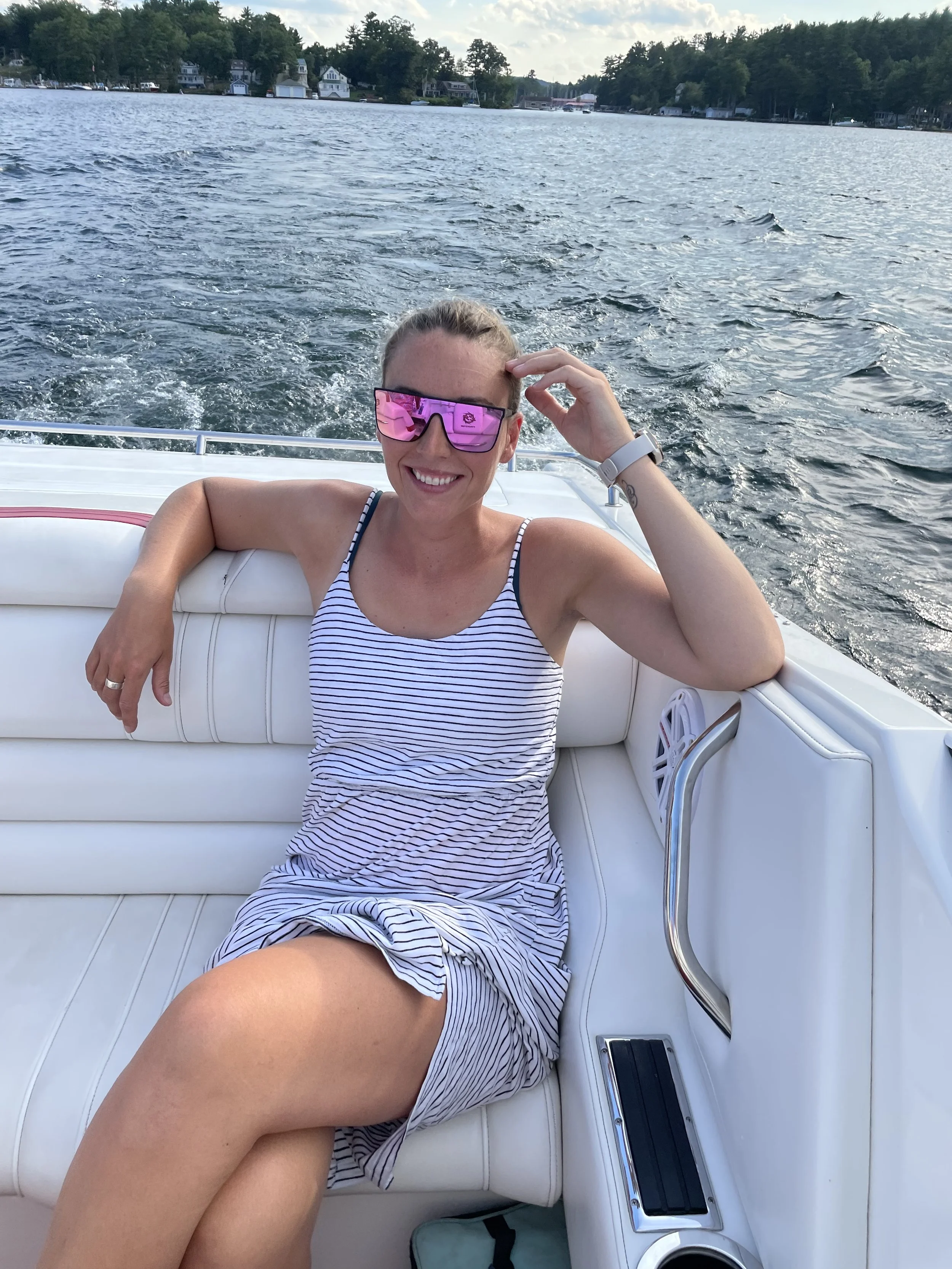 A woman smiling and wearing sunglasses, relaxing on a white boat with a lake and houses in the background.