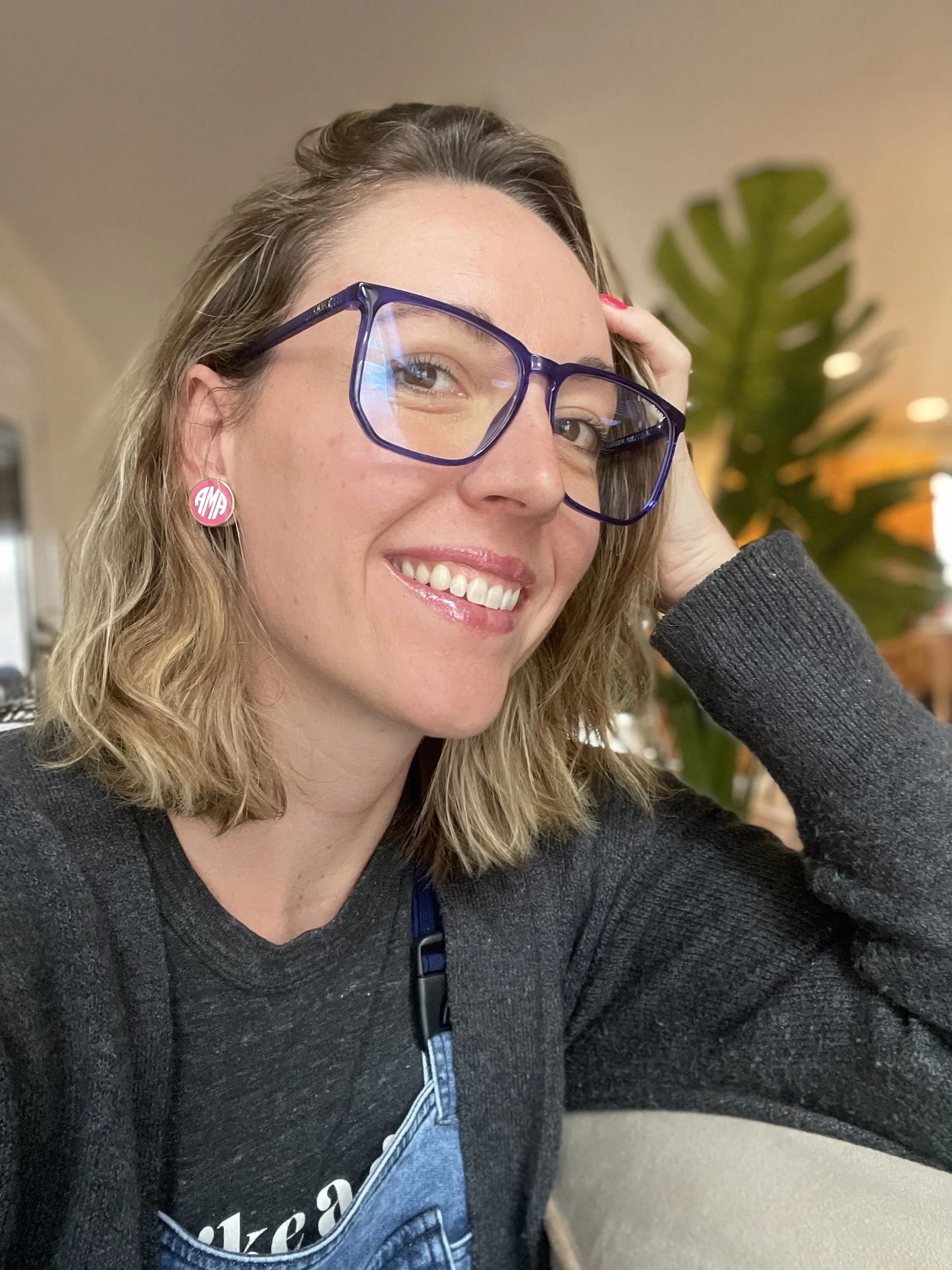 A woman with wavy blonde hair and glasses smiling at the camera, wearing a gray sweater and large pink earrings with 'M' on them, seated indoors with a large green plant in the background.