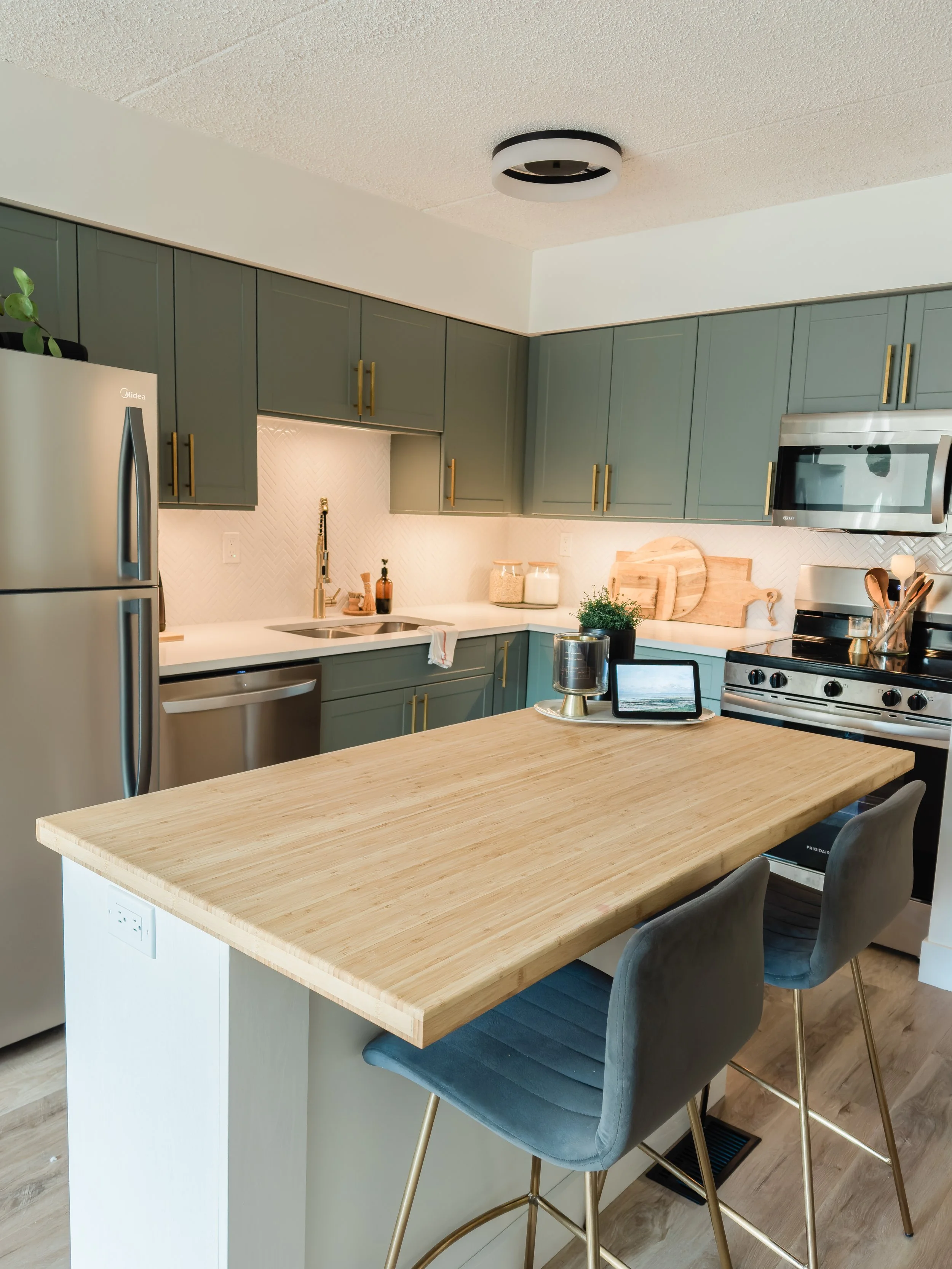 Modern kitchen with green cabinets, wooden countertop, stainless steel appliances, and two high chairs.