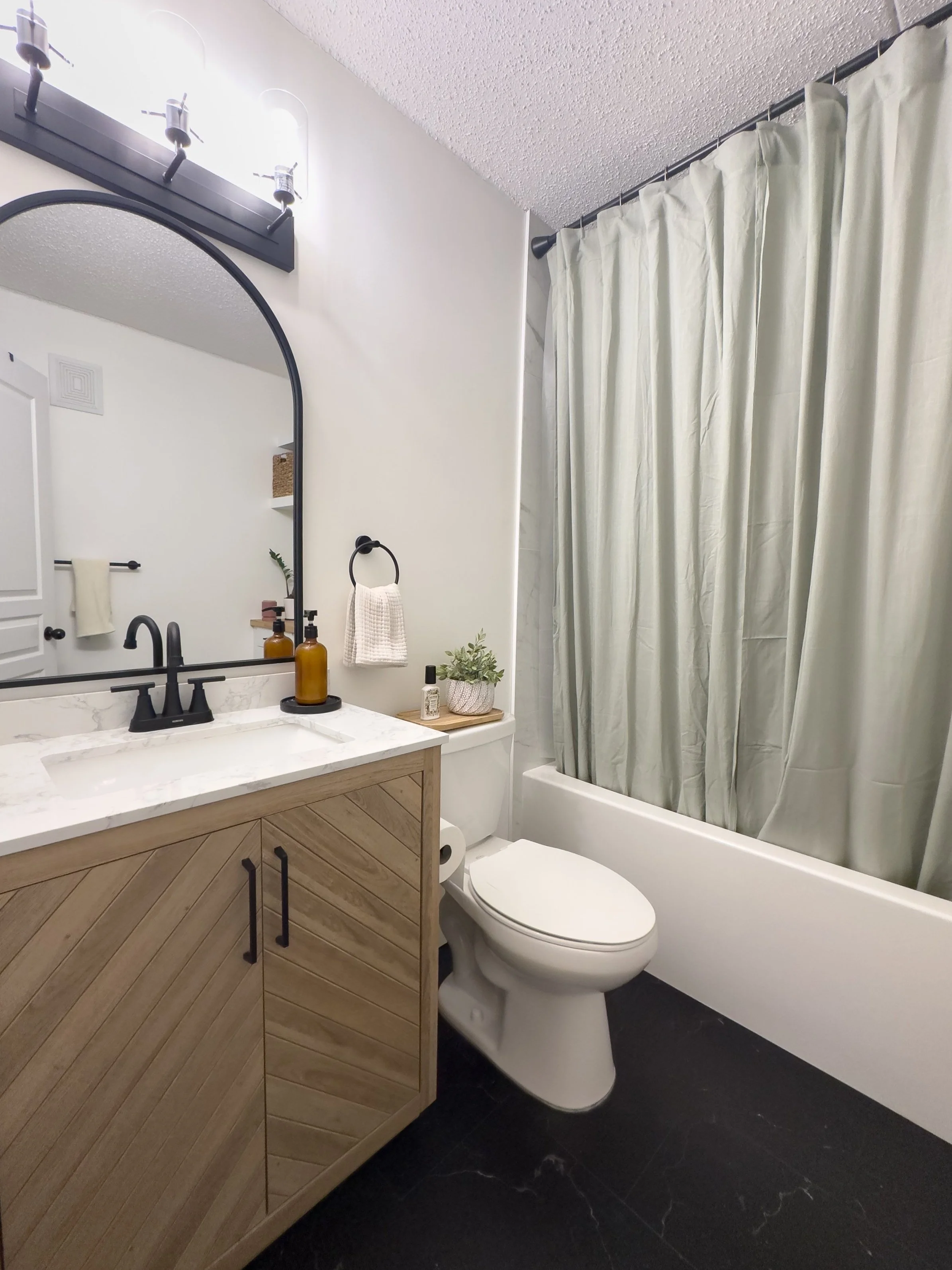 Modern bathroom with wood vanity, marble countertop, oval mirror, black faucet, toilet, bathtub with white shower curtain, dark floor tiles, and wall-mounted lighting.