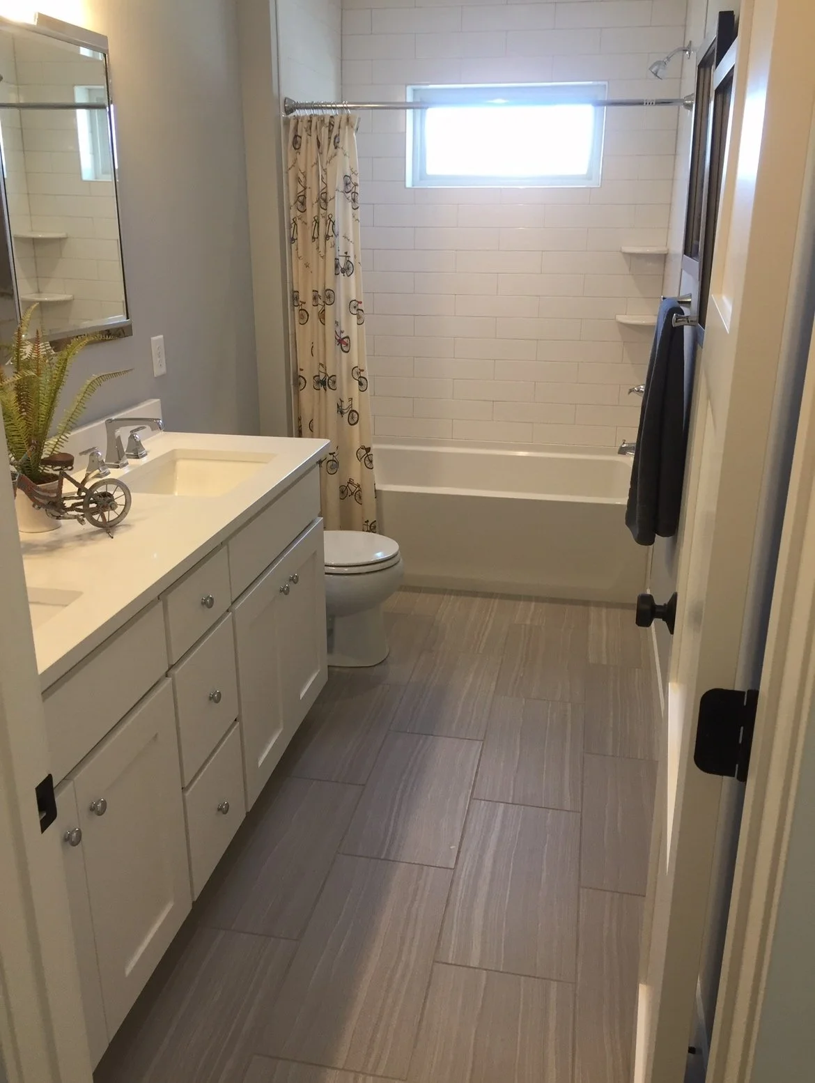 Bathroom with white vanity, a mirror, a toilet, a bathtub with a bicycle-themed shower curtain, and a small window. Tiled floor and shelves in the shower area.