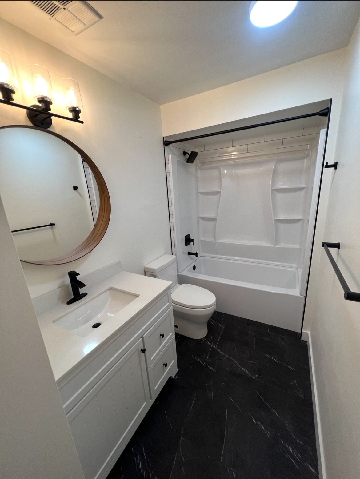 A modern bathroom featuring a white vanity with a sink, black fixtures, a large round mirror, a toilet, and a bathtub with a shower curtain rod, black fixtures, and a built-in shower niche. The floor is black with white veining, and there are black t