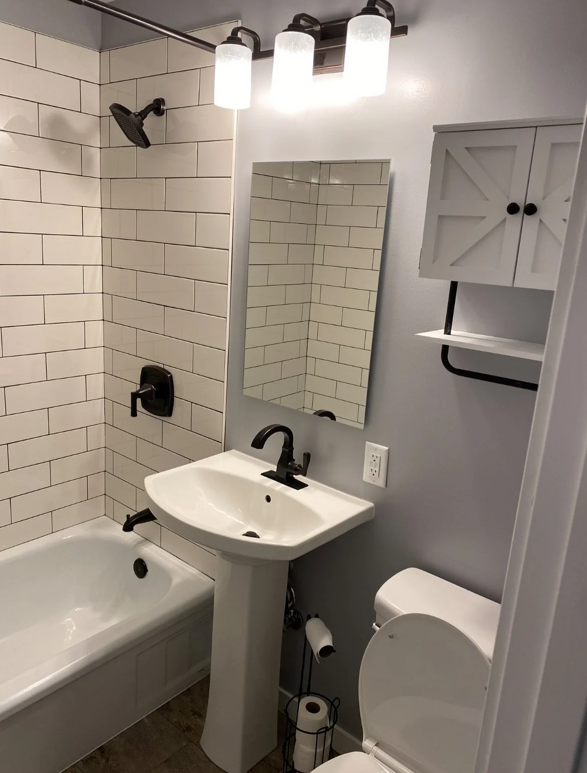 Small bathroom with a bathtub, a pedestal sink with a black faucet, a rectangular mirror, a wall-mounted cabinet, a toilet, and a black fixture above the bathtub.