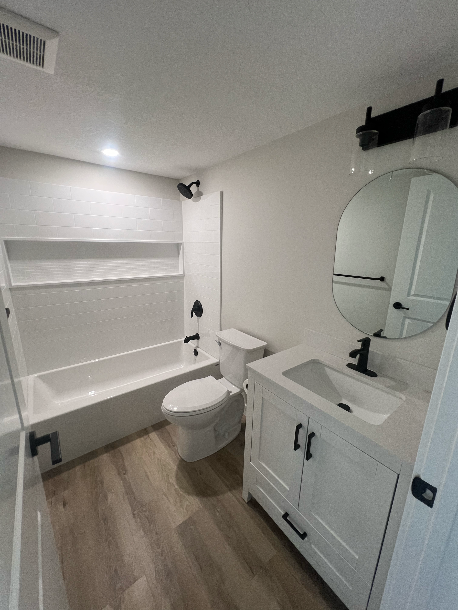 White bathroom with a vanity, round mirror, and toilet, black fixtures, and wood flooring.