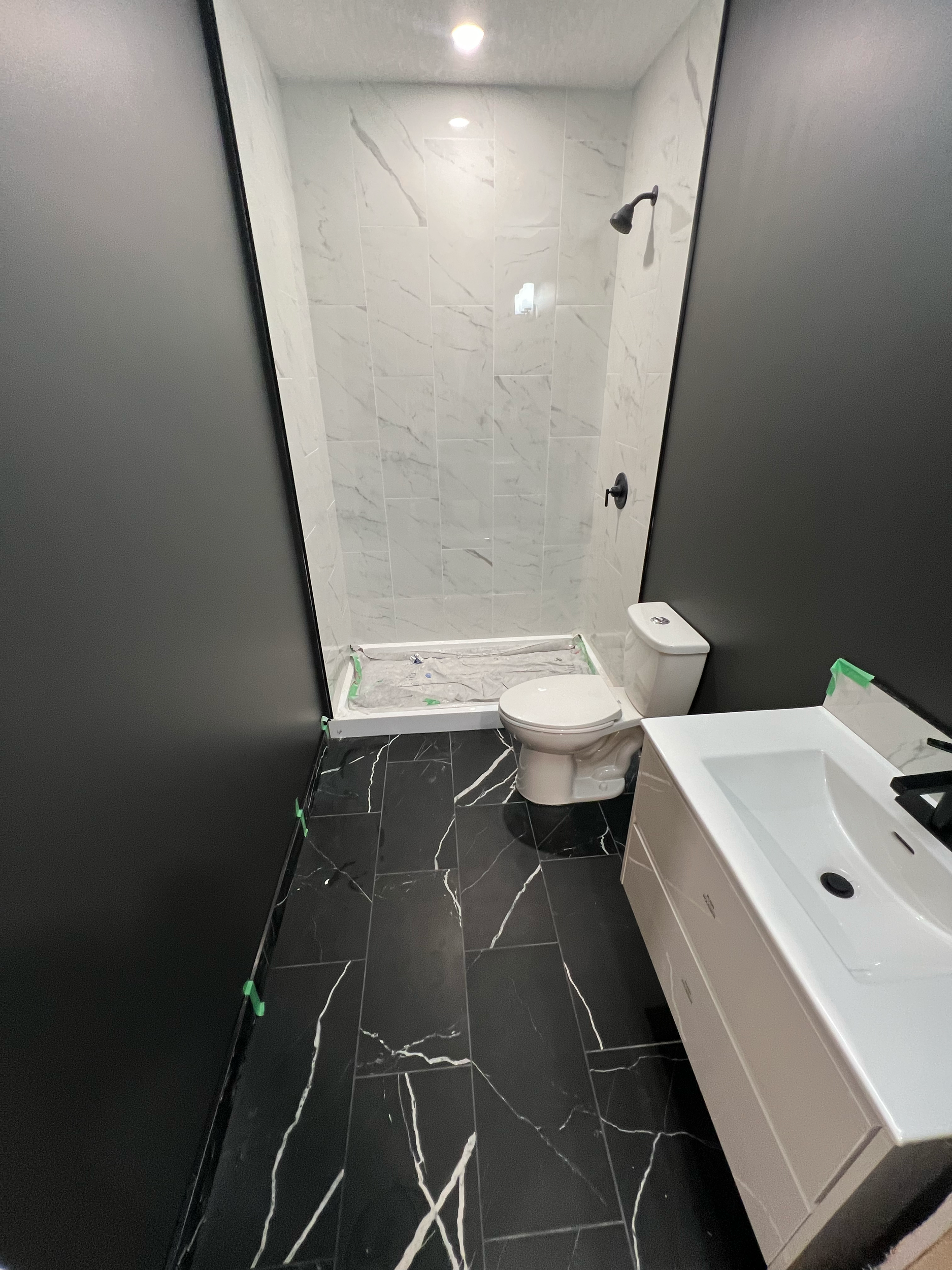 Partially renovated bathroom with black and white marble tile flooring, white marble wall tiles in the shower area, a white toilet, a white sink cabinet, a partially installed toilet, and a dark ceiling with two recessed lights and a black showerhead