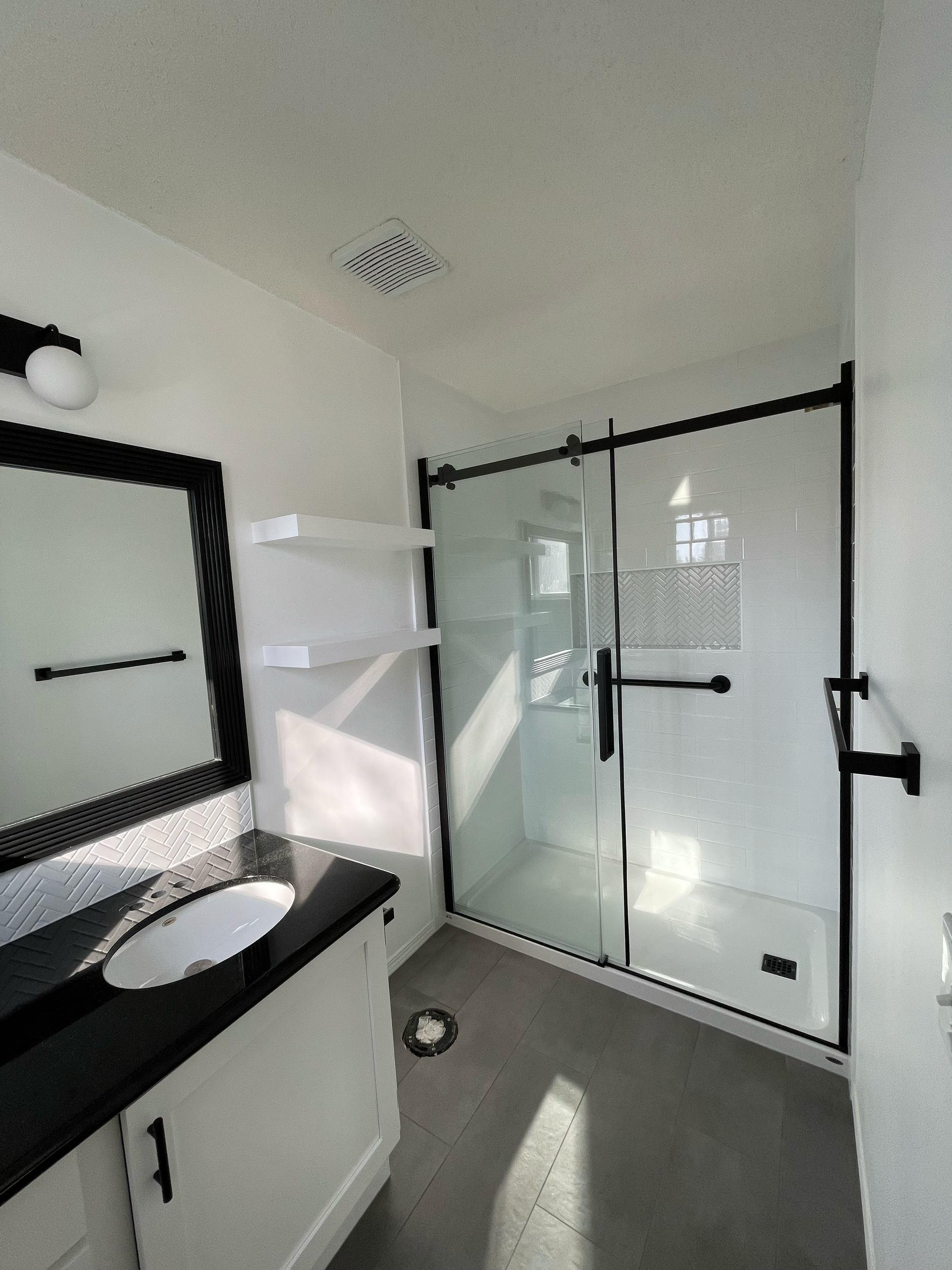 Modern bathroom with a glass shower enclosure, white walls, a black-framed mirror, a black vanity with a white sink, and gray tile flooring.
