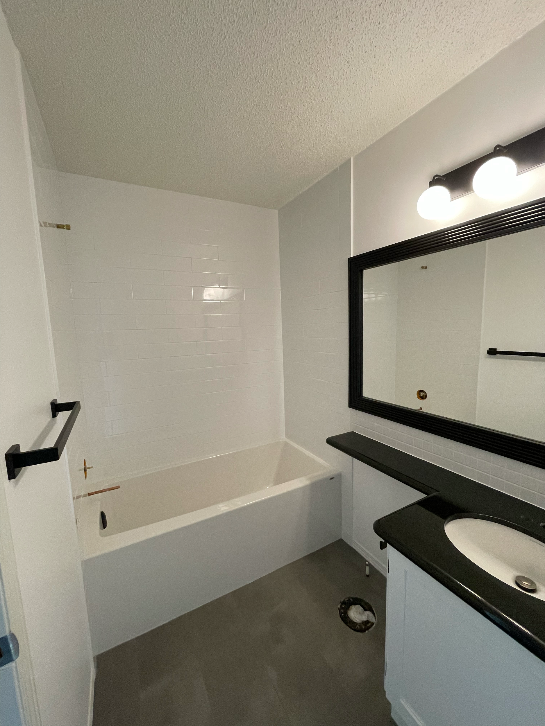 Bathroom with white tiled wall, black vanity with a white sink, large mirror, and a wall-mounted light fixture with three round bulbs.