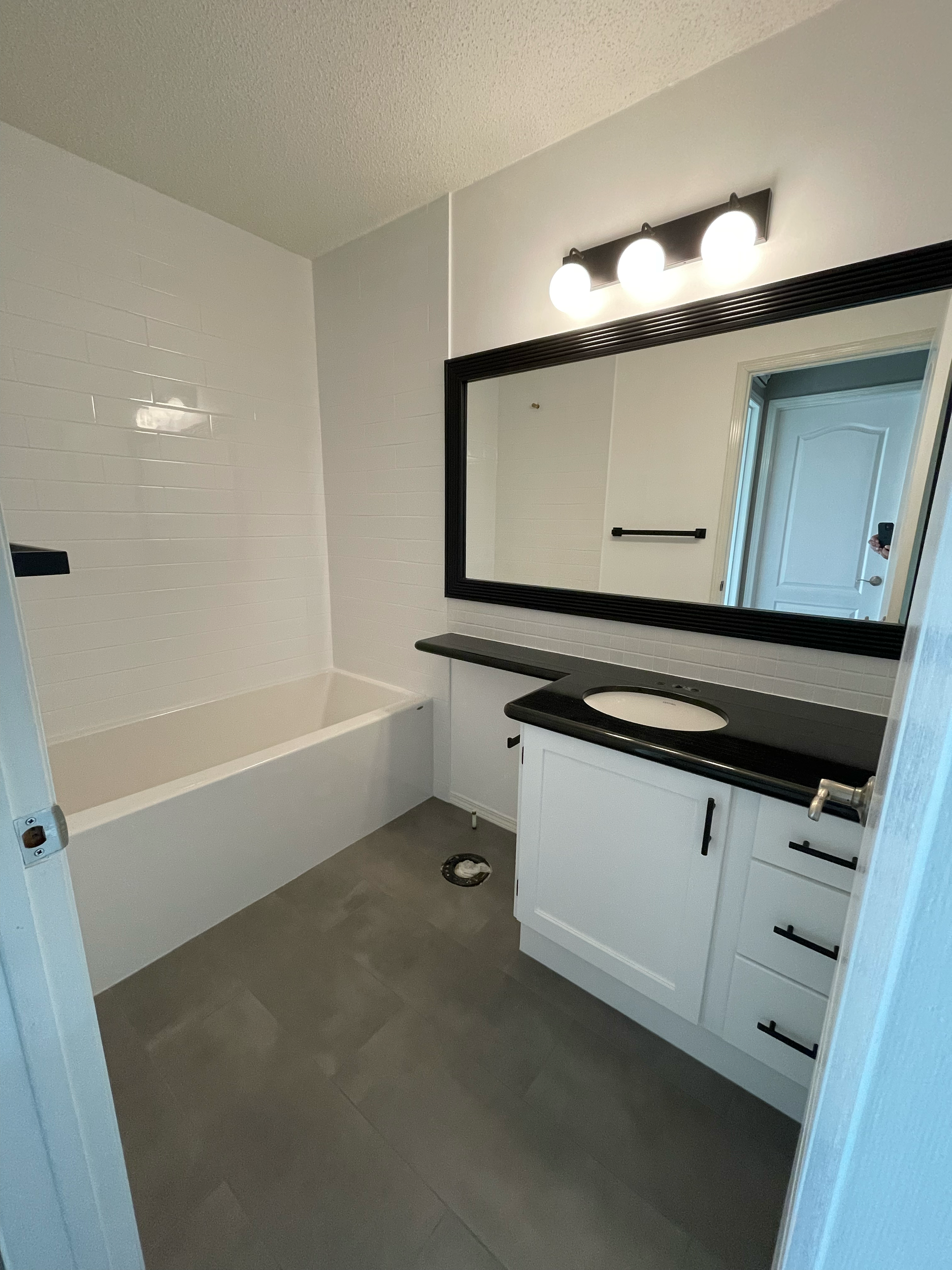 A bathroom with white cabinets, a black countertop, a large mirror with black trim, and a three-bulb light fixture. There is a door reflected in the mirror, and the wall behind the mirror has a black towel bar. The floor is dark gray tile.