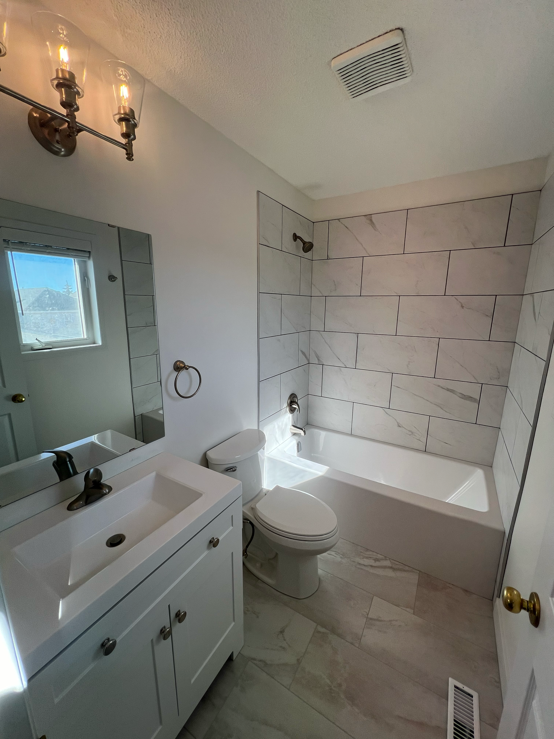 A bathroom with white walls, a toilet, a sink with a white vanity, a large mirror, a small window, a frame, a towel ring, and a light fixture with three Edison bulbs. The bathtub area is tiled with large beige and white marble tiles, and the floor ha