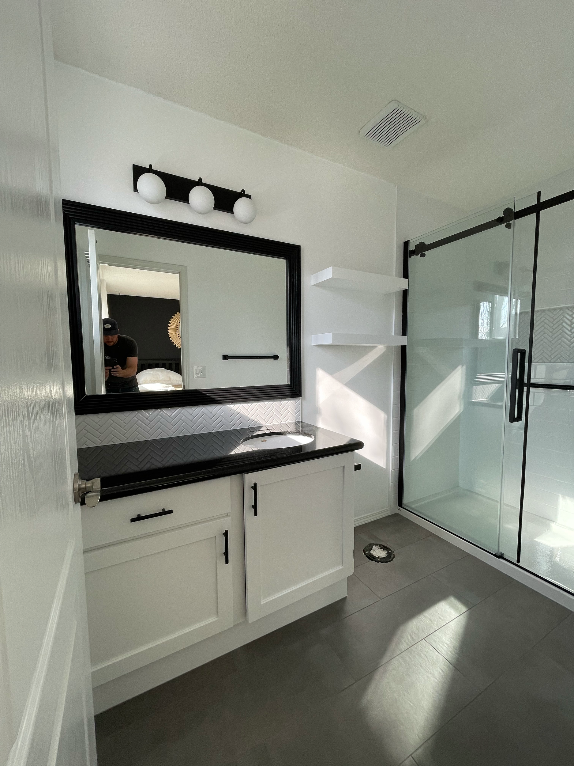 Bathroom with black-framed mirror, white vanity with black handles, black countertop, gray tile floor, glass shower door, and white wall shelf. Reflection shows a person taking the photo.