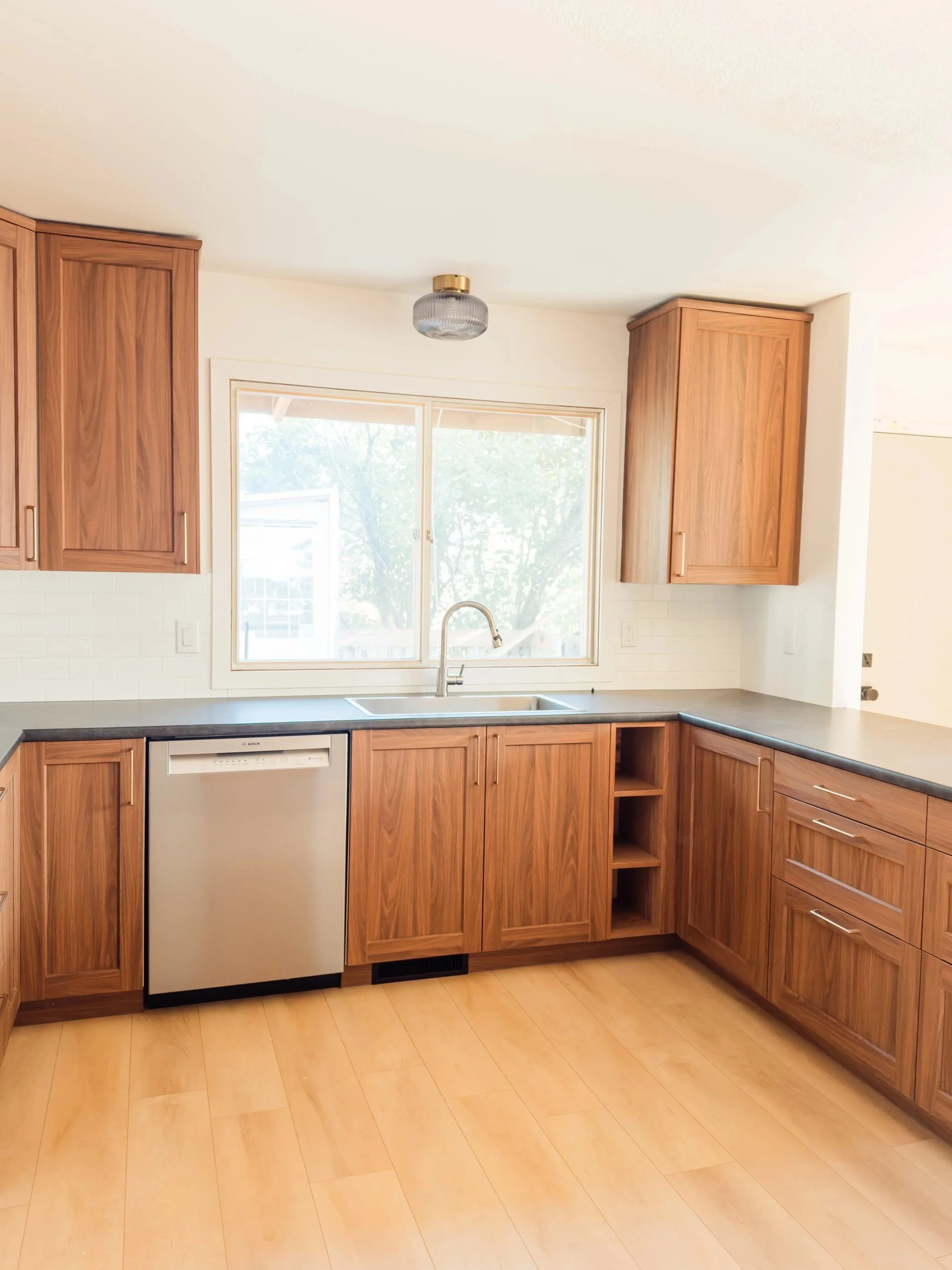 Empty kitchen with wooden cabinets, black countertop, a stainless steel dishwasher, and a window above the sink.