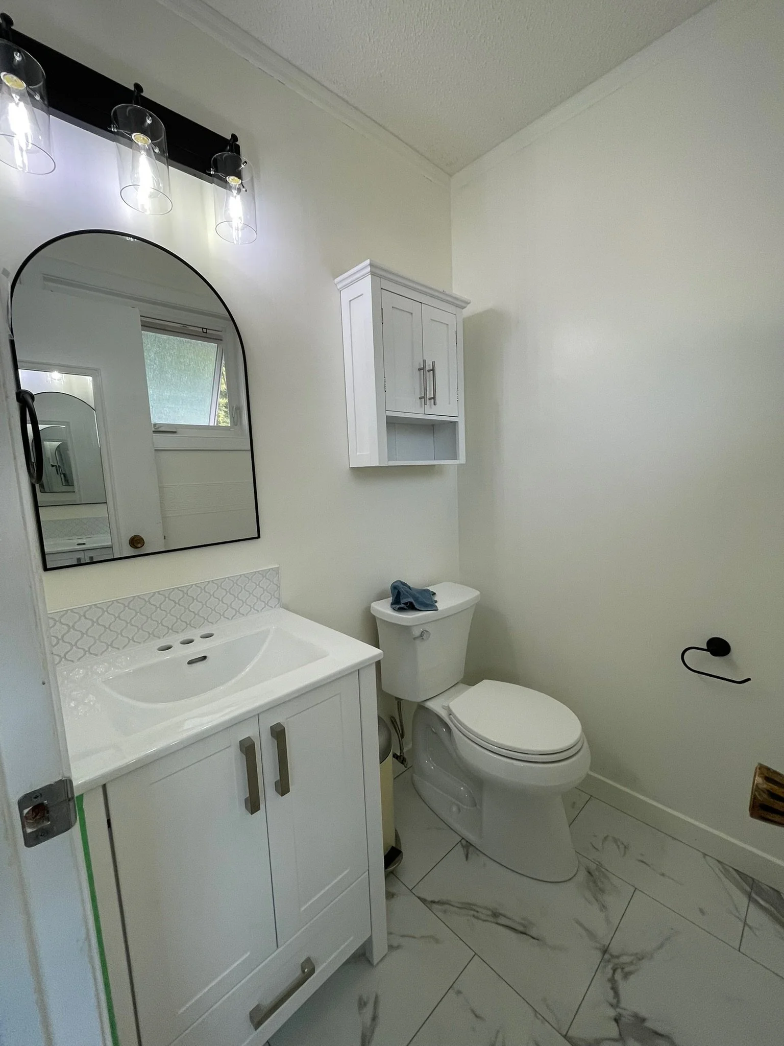 A small bathroom with a white vanity and an arched mirror, a wall-mounted cabinet, a toilet with a blue cloth on the tank, a window, and a marble-patterned floor.