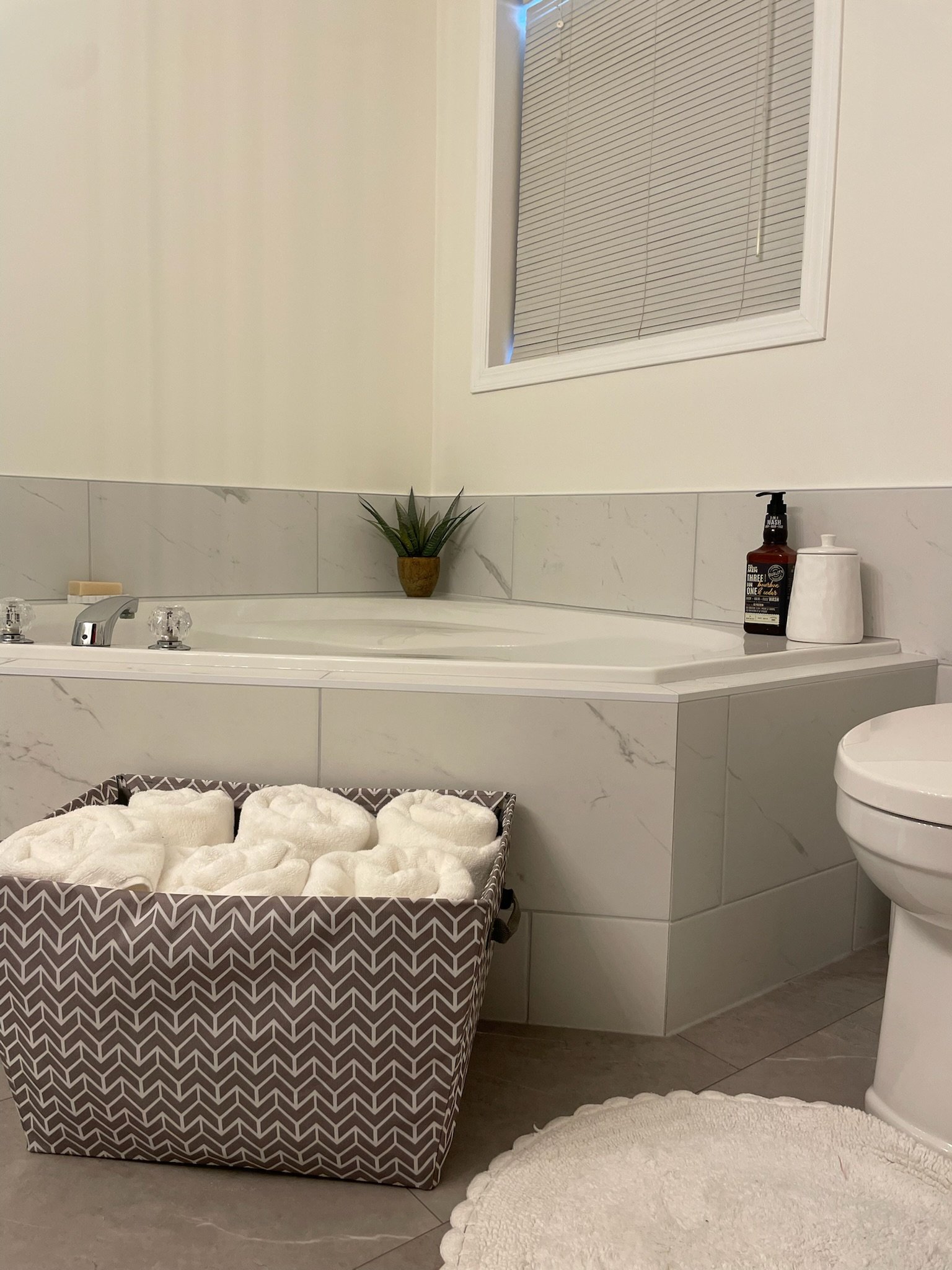 Bathroom with a corner bathtub, a basket of rolled white towels, a potted plant, a toilet, and toiletries on the windowsill.