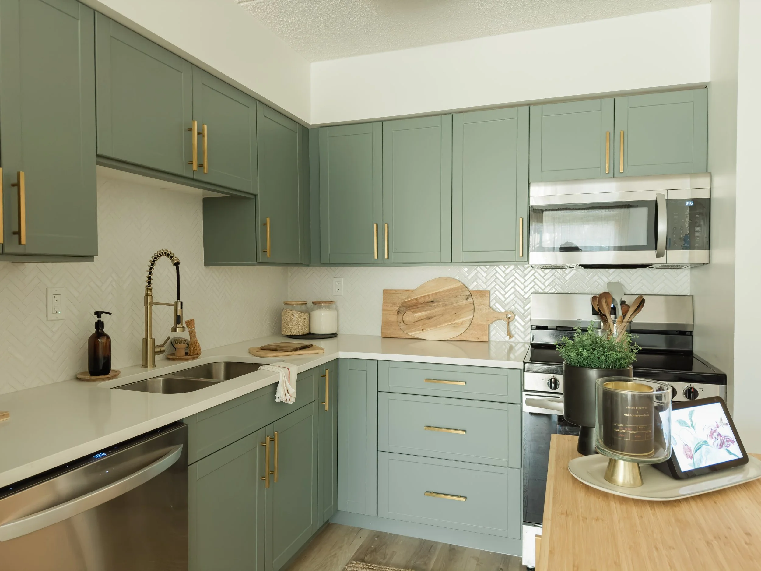 Modern kitchen with green cabinets, brass handles, white countertops, stainless steel appliances, and decorative items like a potted plant and wooden utensils.