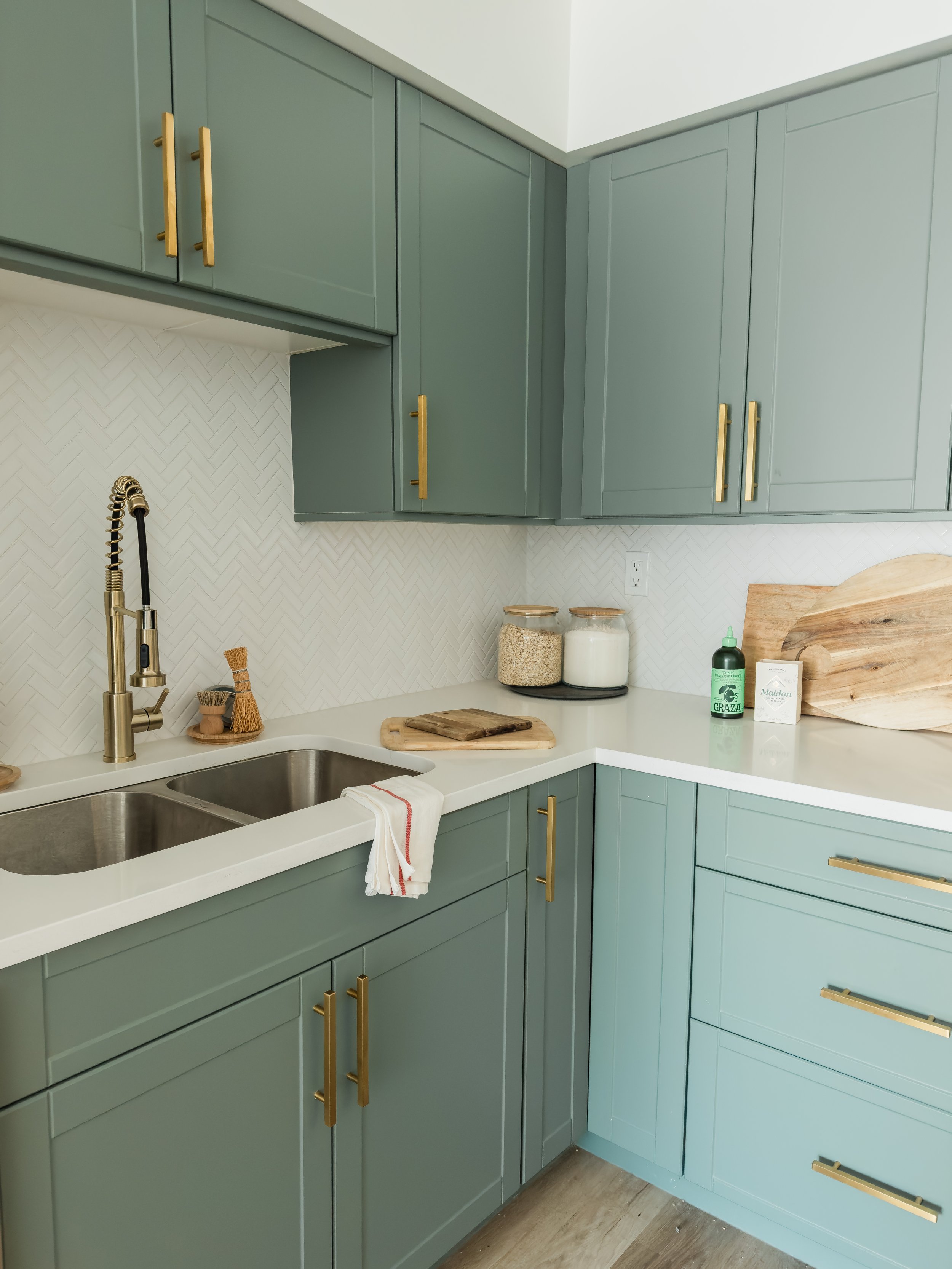 Modern kitchen with green cabinets, gold handles, double stainless steel sink, white countertop, herringbone backsplash, wooden cutting boards, and jars on the counter.