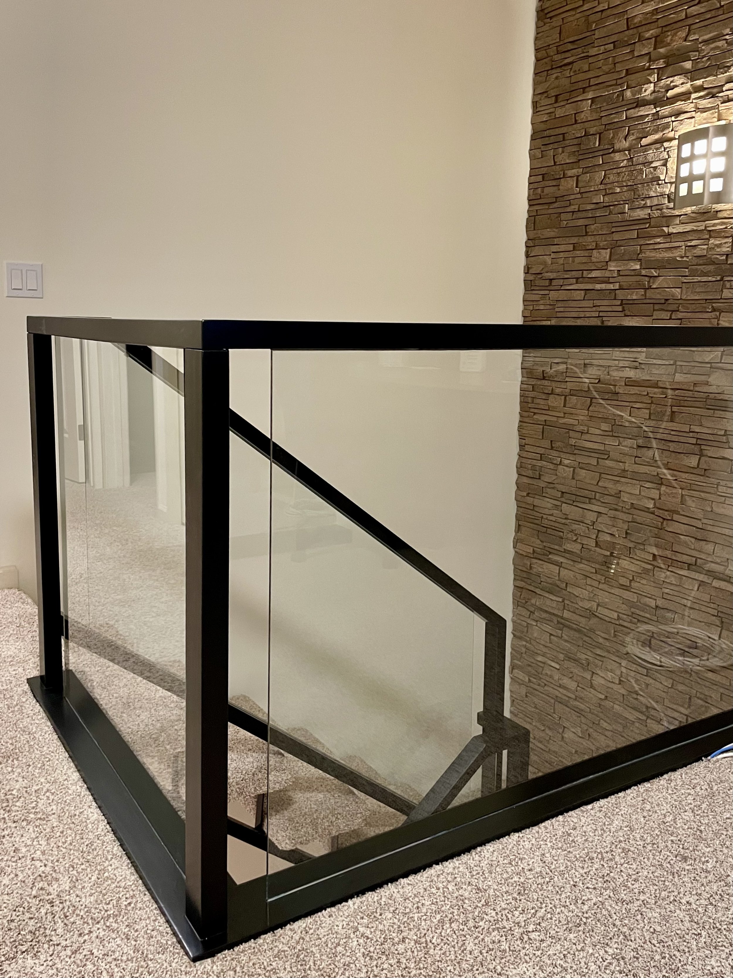 Modern staircase with glass railing and black frame, carpeted flooring, exposed brick wall, and wall-mounted light fixture.