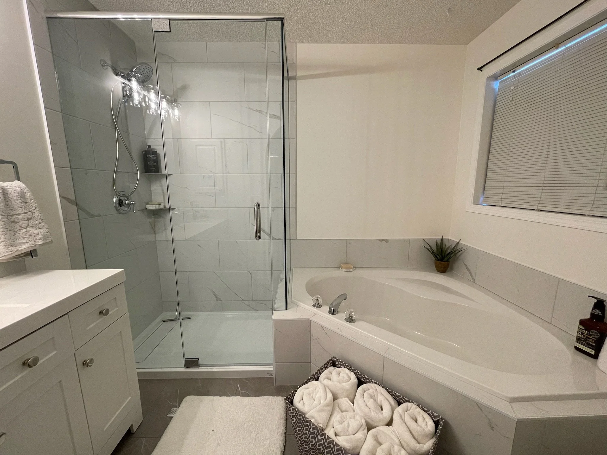 Bathroom with glass shower enclosure, white vanity with towels, a bathtub with rolled towels, a potted plant by the window, and bottles on the bathtub