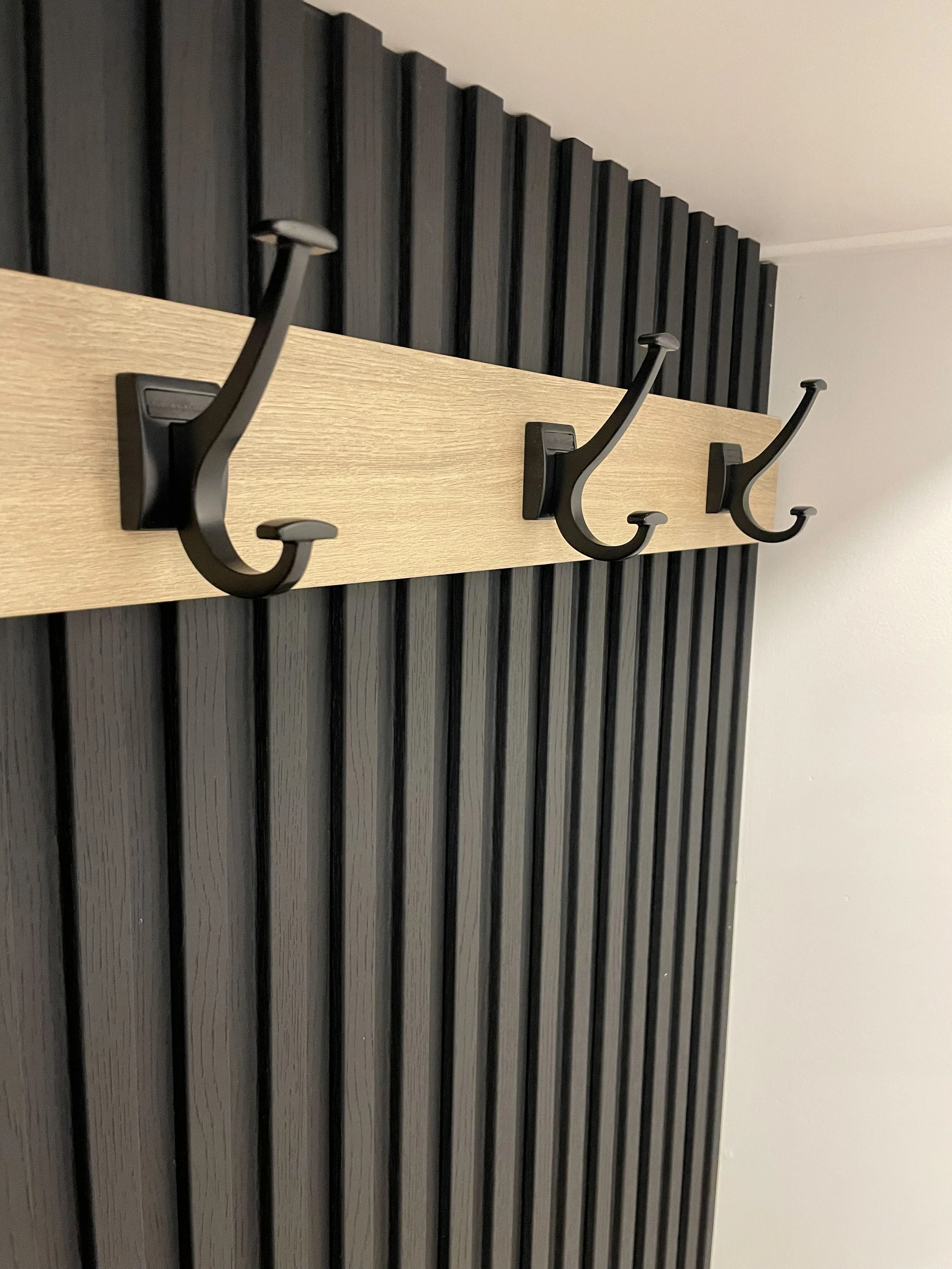Three black coat hooks mounted on a light wood panel against a black slatted wall.