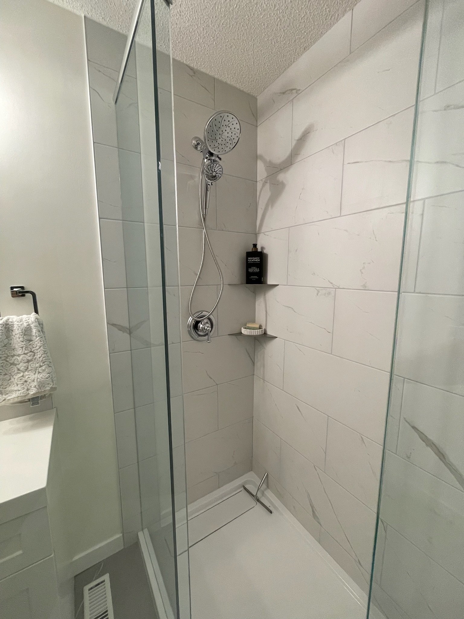 A clean, modern shower with a glass door, white marble walls, a chrome showerhead and handheld shower, a small corner shelf with toiletries, and a white floor.