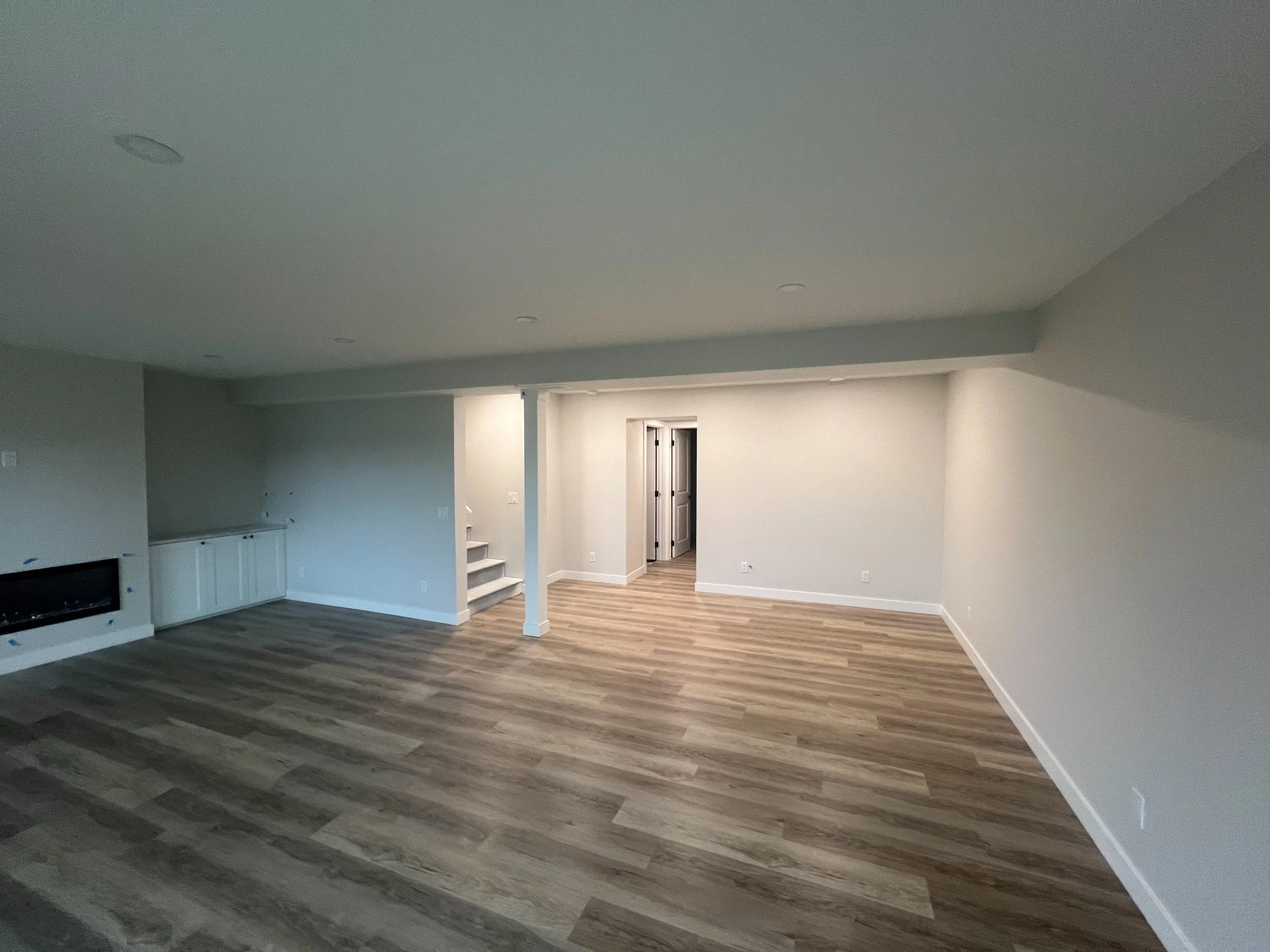 Empty room with wooden flooring, recessed lighting, and light gray walls; a fireplace and built-in cabinets are visible on one side. Stairs lead to a doorway.