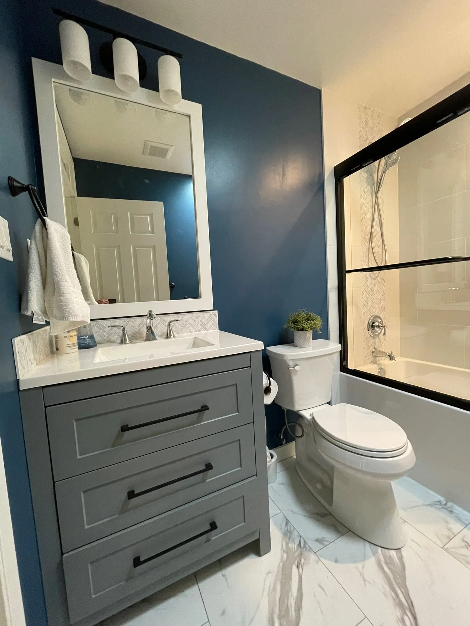 A modern bathroom with a gray vanity, white sink, and a large mirror. There are three white lights above the mirror. To the right, there is a white toilet with a small potted plant on top. Next to the toilet is a glass shower enclosure with a black f