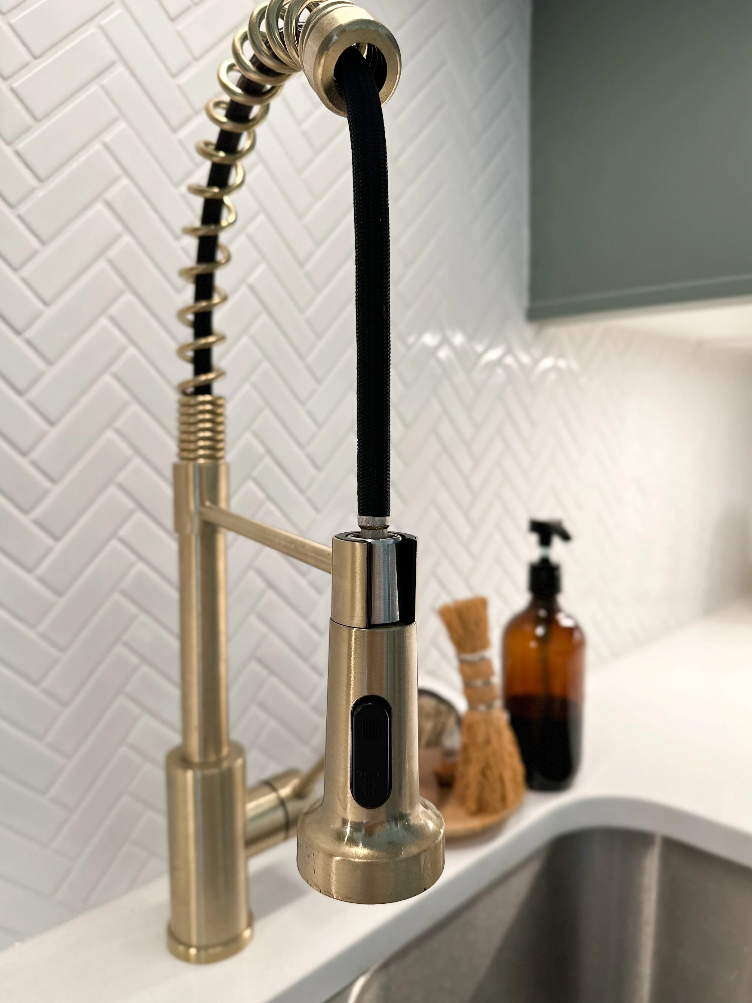 Modern kitchen faucet with pull-down sprayer in front of a herringbone tile backsplash, next to a soap dispenser and cleaning brushes.