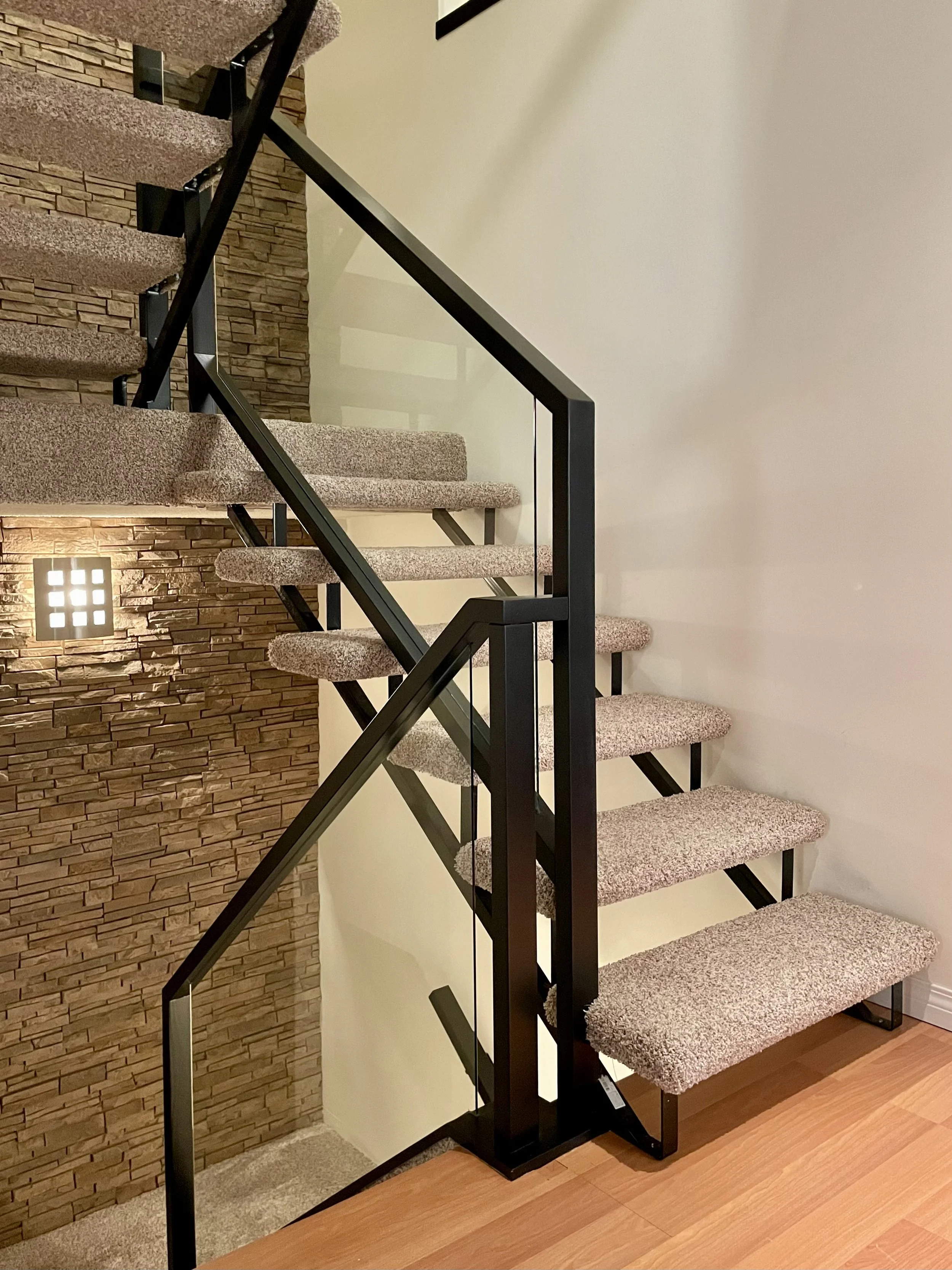 Modern staircase with carpeted steps and metal railings, featuring a stone accent wall and a wooden floor.