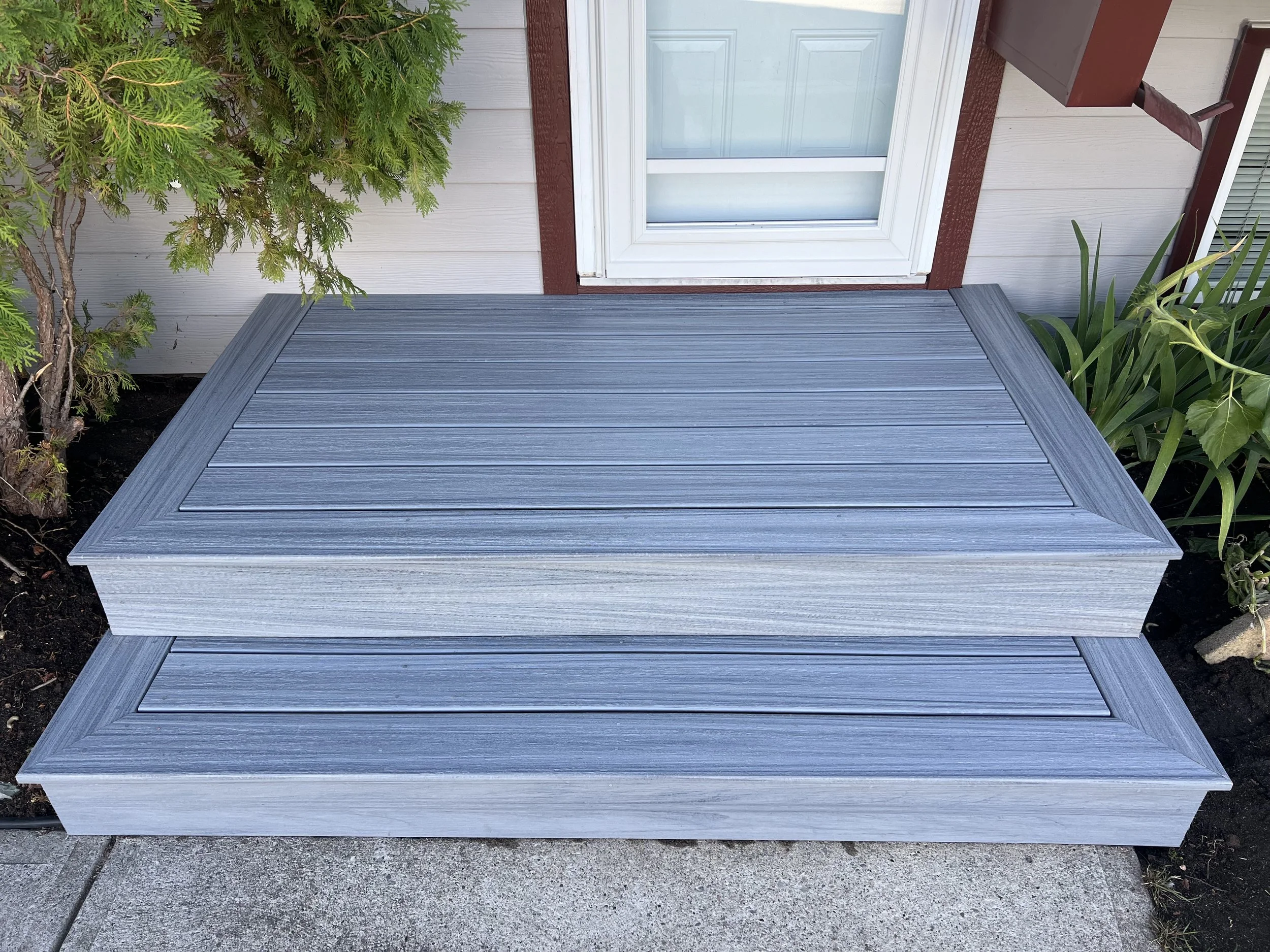Gray composite decking steps leading to a house door, surrounded by greenery.