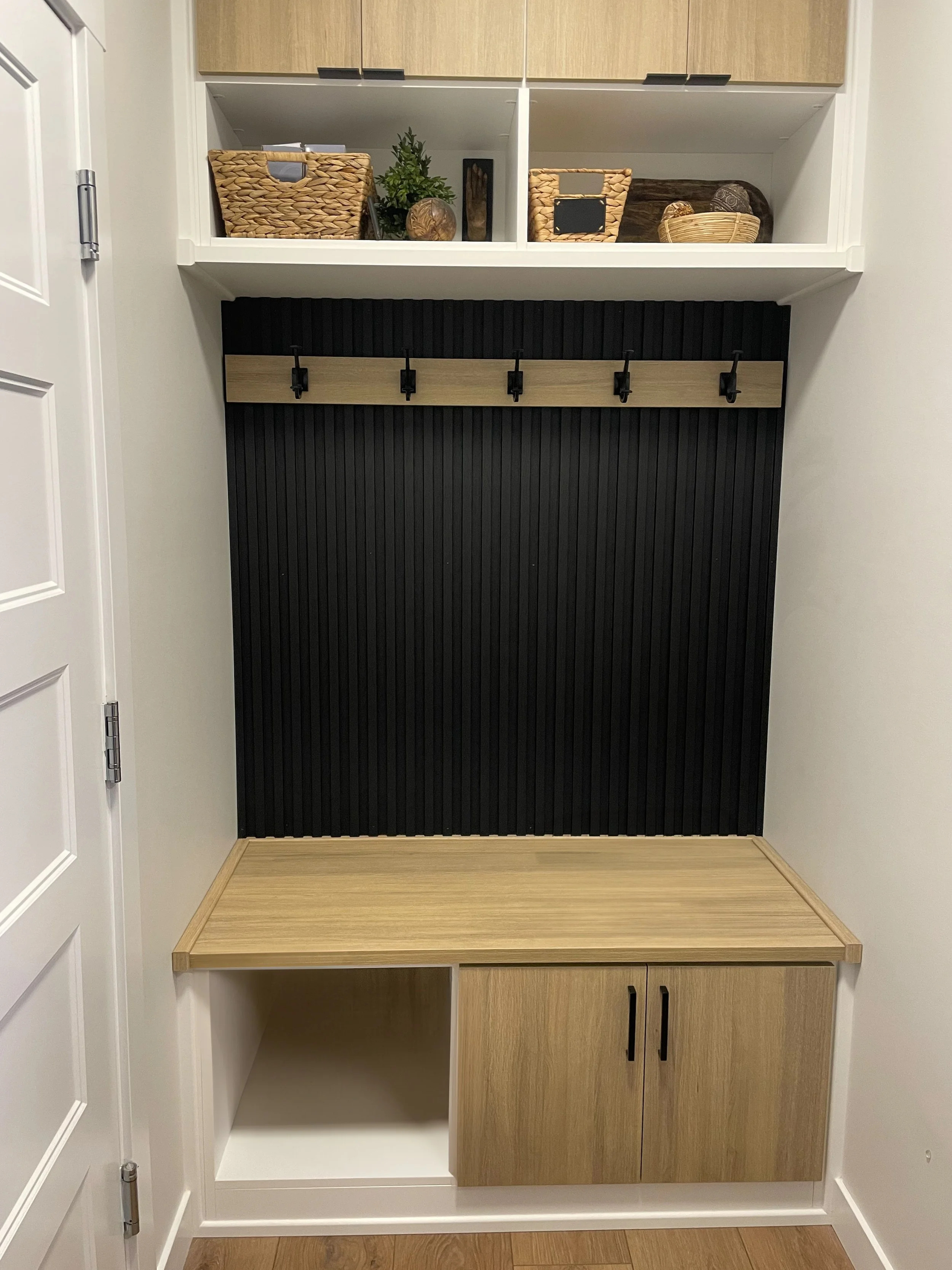 Modern entryway with wooden bench, black panel wall, coat hooks, overhead shelves with wicker baskets and decor items.