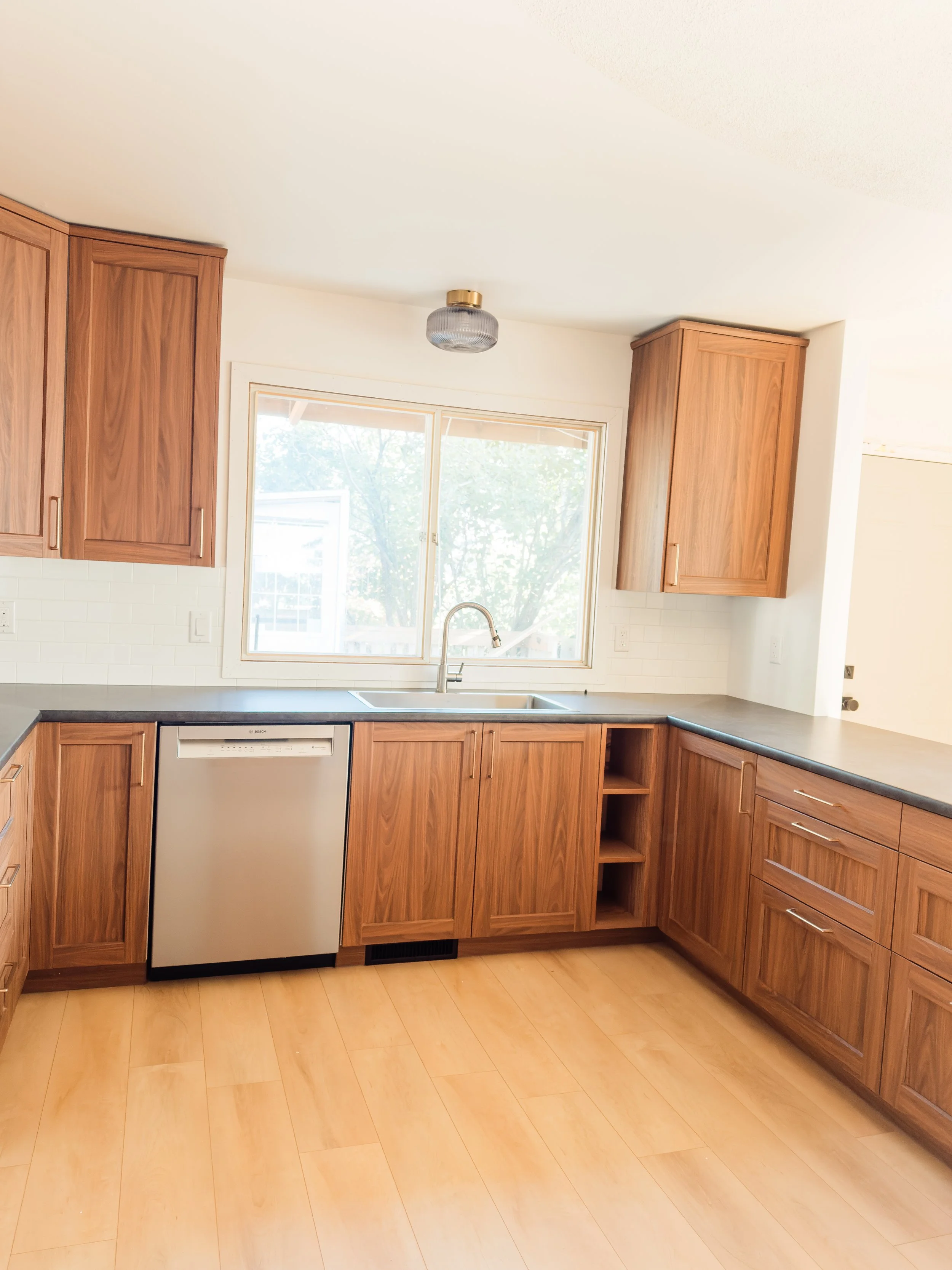 Empty kitchen with wooden cabinets, black countertop, a stainless steel dishwasher, and a window above the sink.