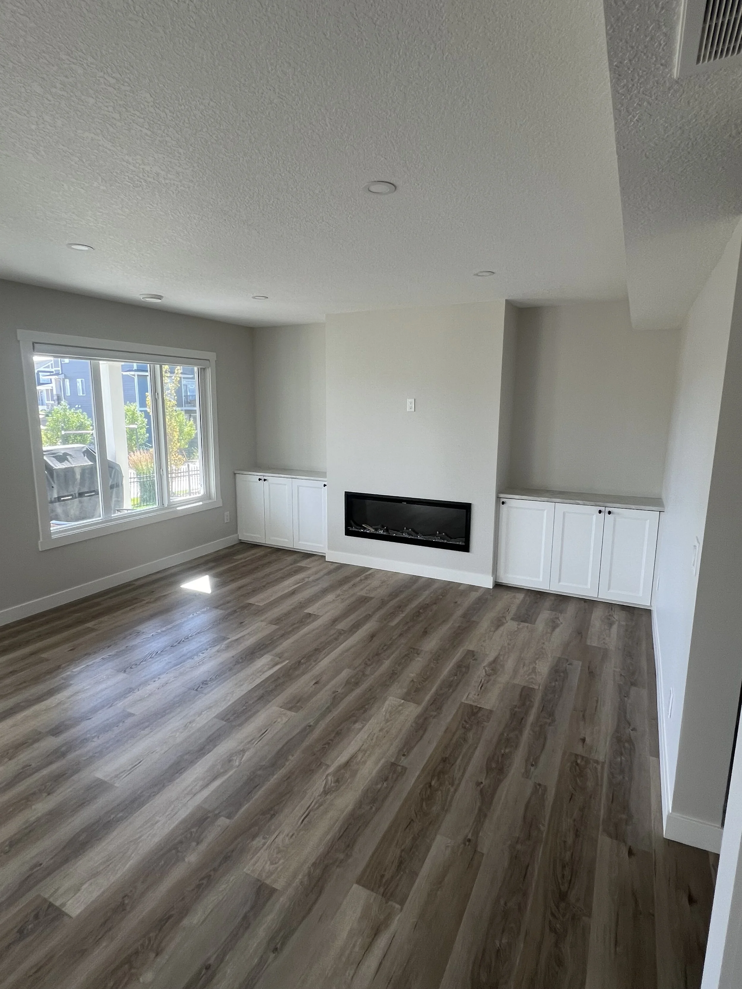 Bright, empty living room with hardwood floors, recessed lighting, a modern fireplace, and built-in cabinets beneath a large window.