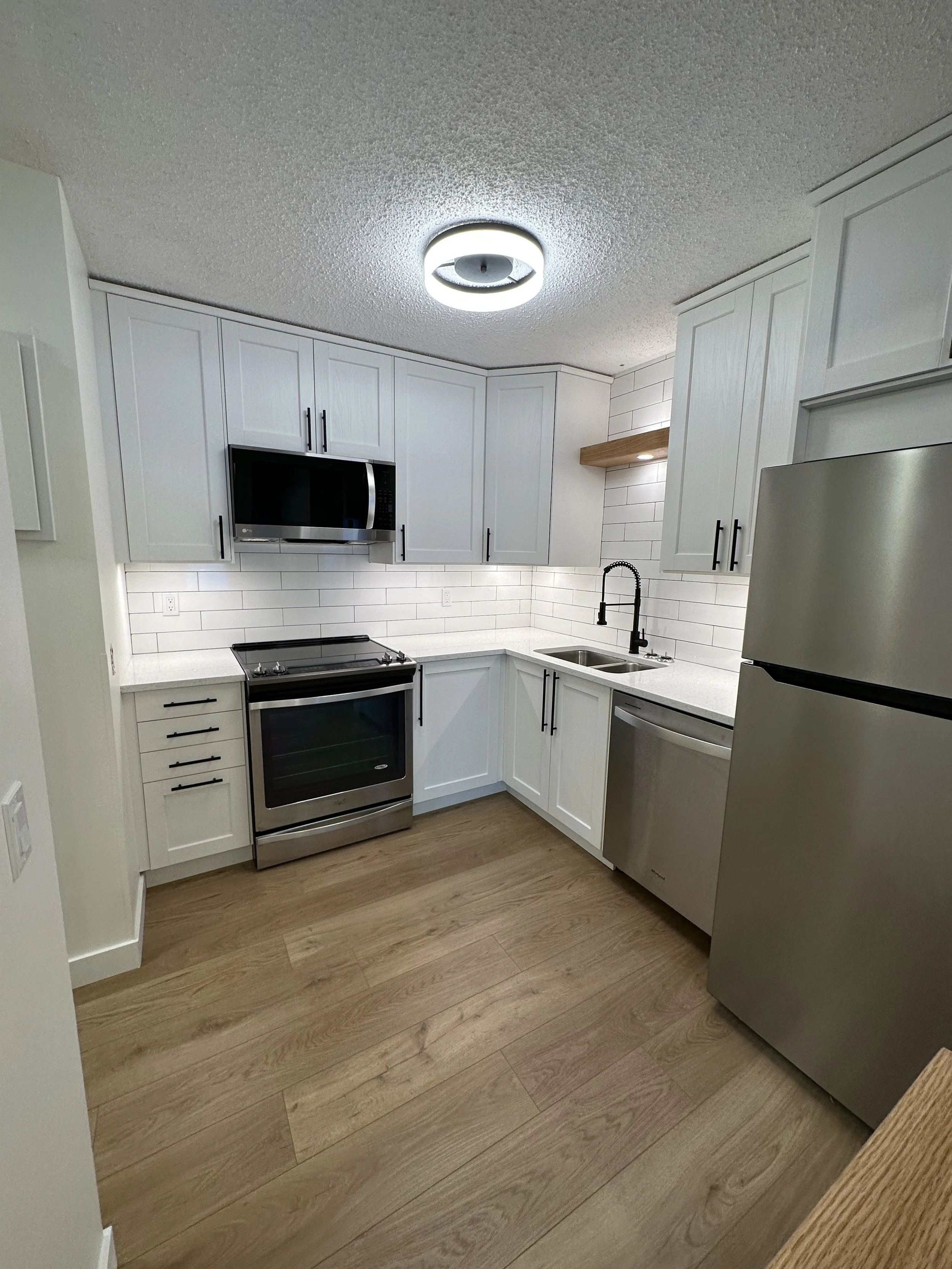 Modern kitchen with stainless steel appliances, white cabinets, and wooden flooring. Features a microwave, oven, refrigerator, sink, and under-cabinet lighting.