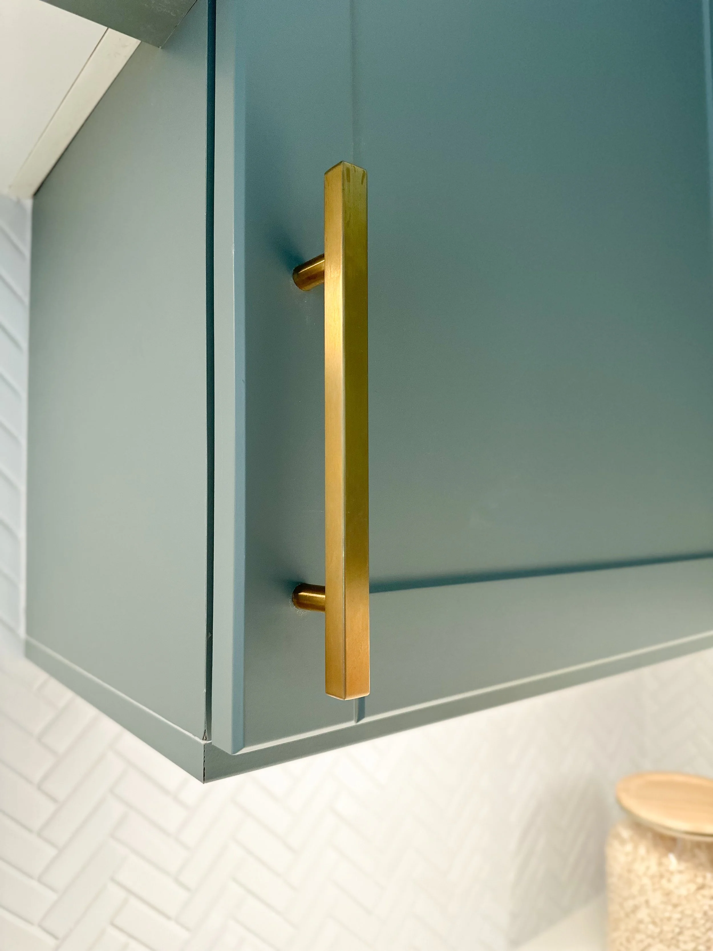 Close-up of a blue kitchen cabinet with a gold handle, white herringbone backsplash, and a container with a wooden lid.