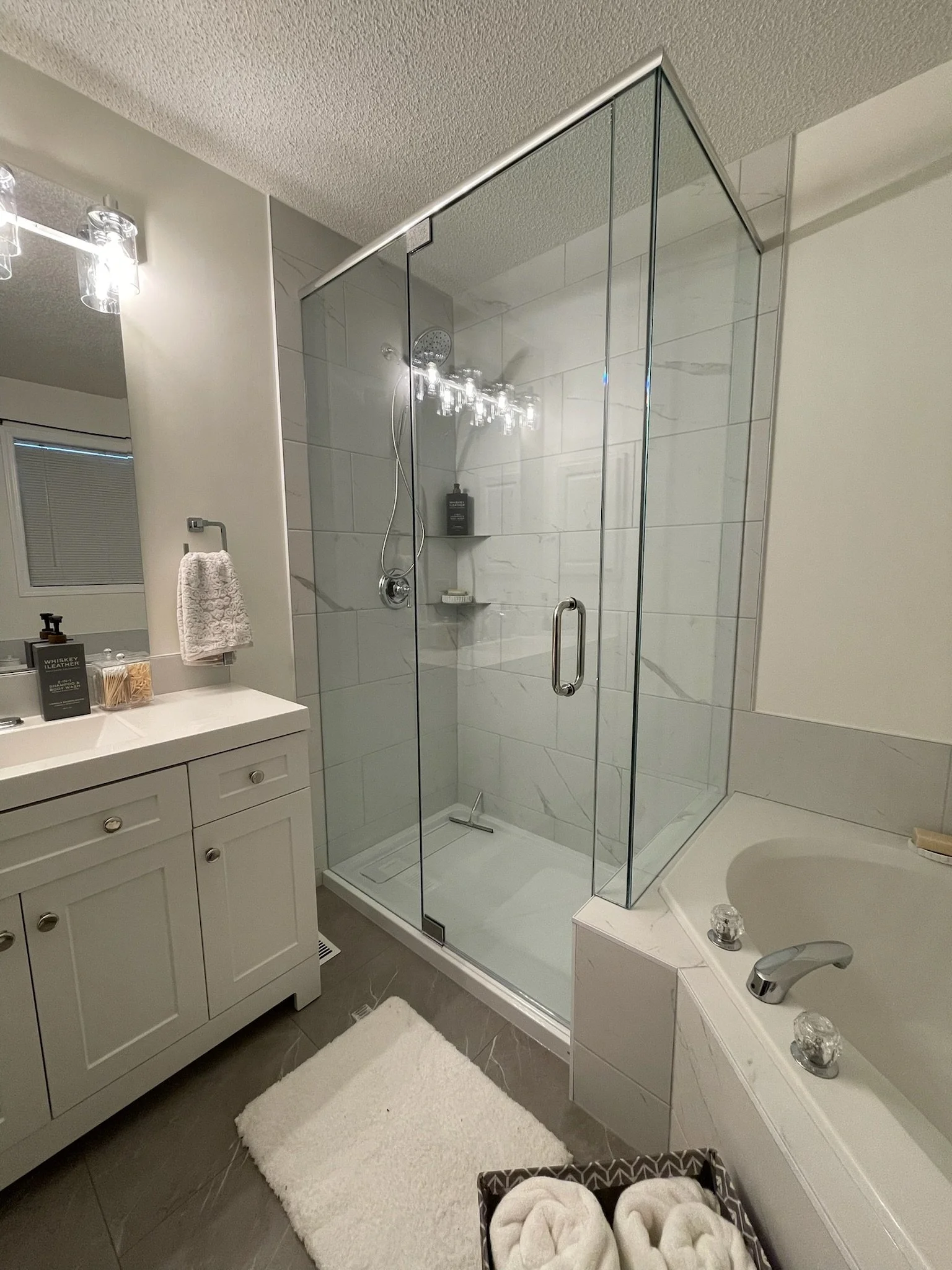 Bathroom with glass-enclosed shower, white vanity with mirror, bathtub with towels, and a white rug on brown tiled floor.