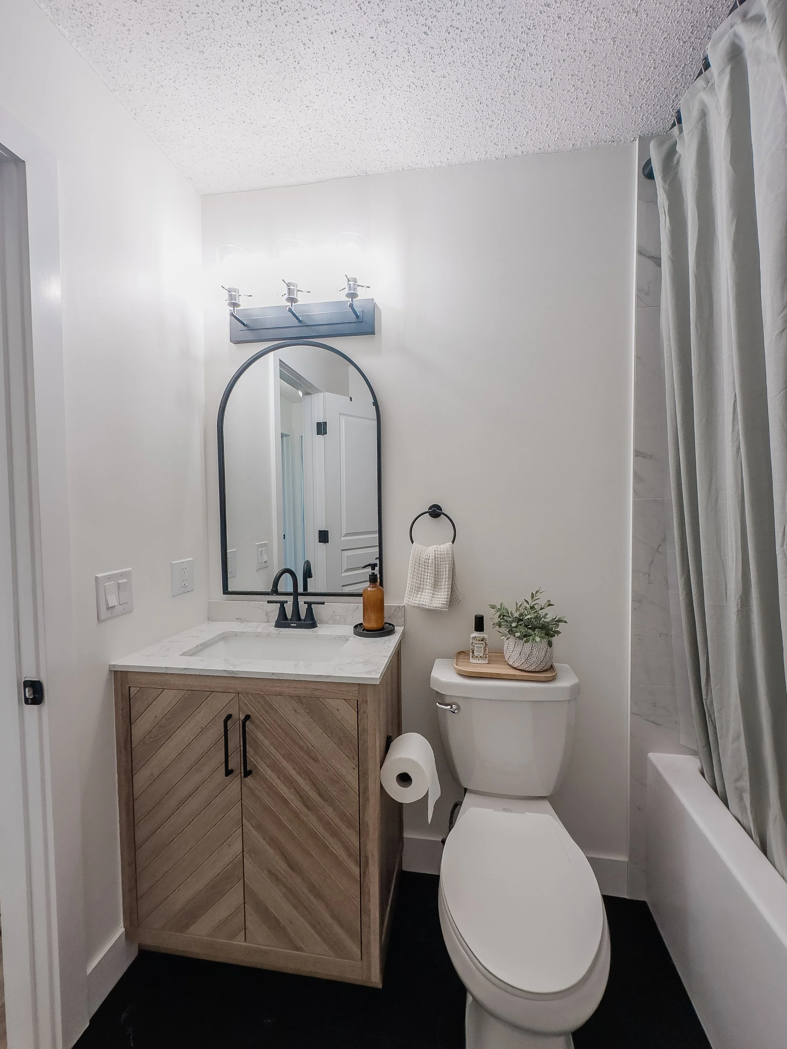 Modern bathroom with a small vanity featuring a chevron-patterned cabinet, white marble countertop, and black faucet. Above it is an oval mirror with a three-light fixture. Toilet with a plant and decor on the tank sits adjacent, alongside a bathtub 