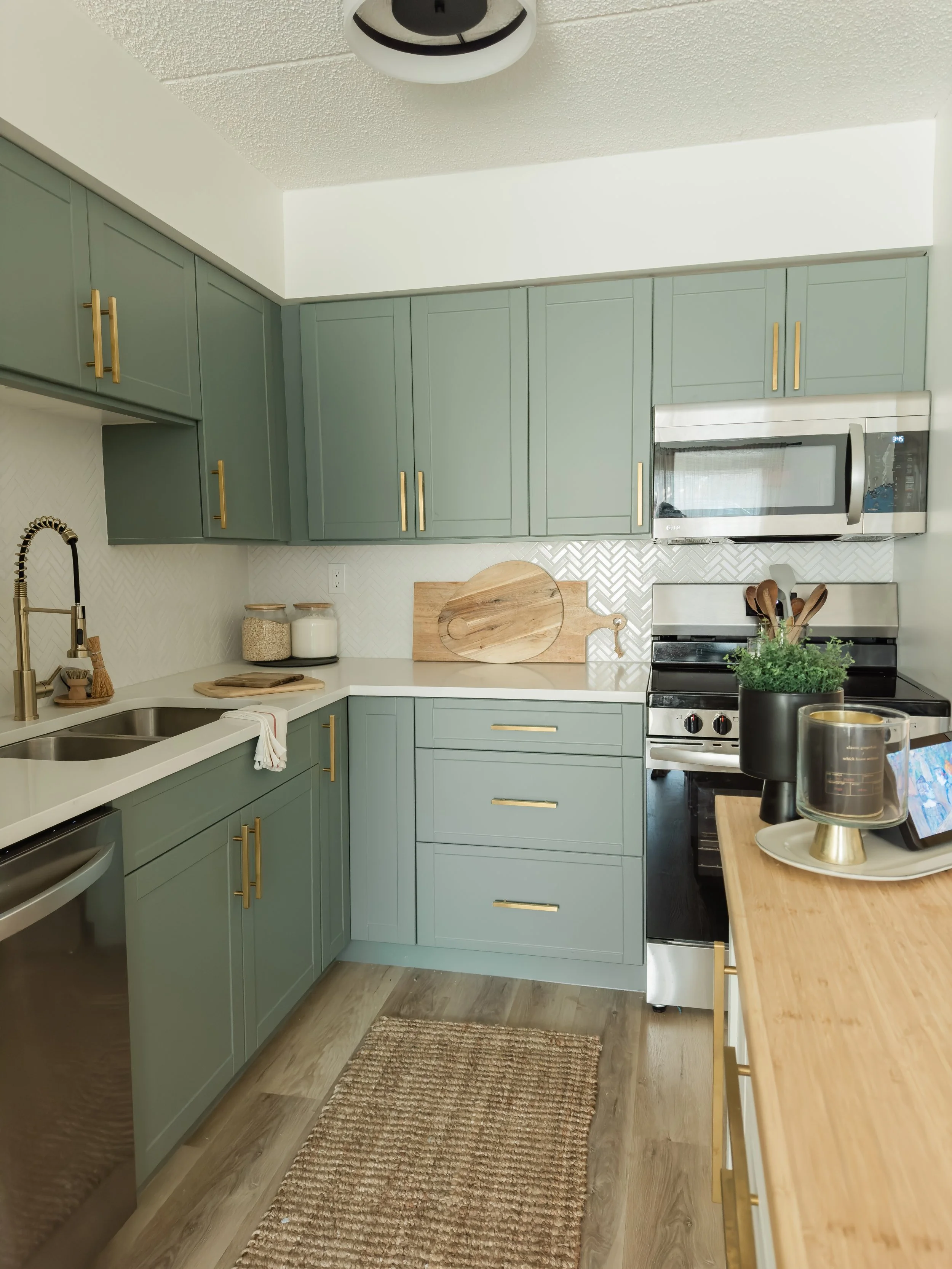 Modern kitchen with green cabinets, gold handles, stainless steel appliances, wooden countertop, and a woven rug on hardwood floor.