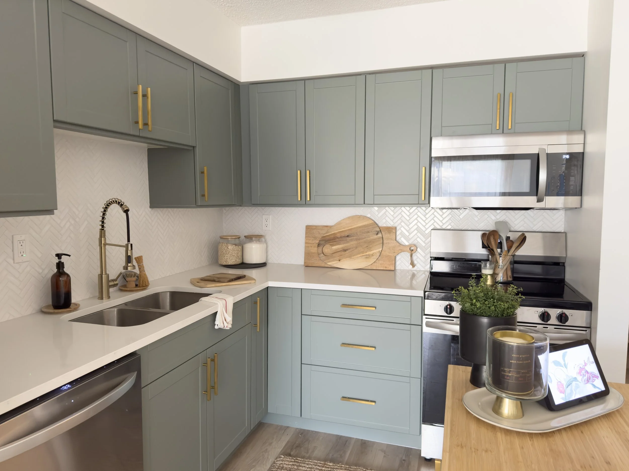 Modern kitchen with green cabinets, white countertops, stainless steel appliances, and gold hardware. A double sink and decorative items, including cutting boards and plants, are present.