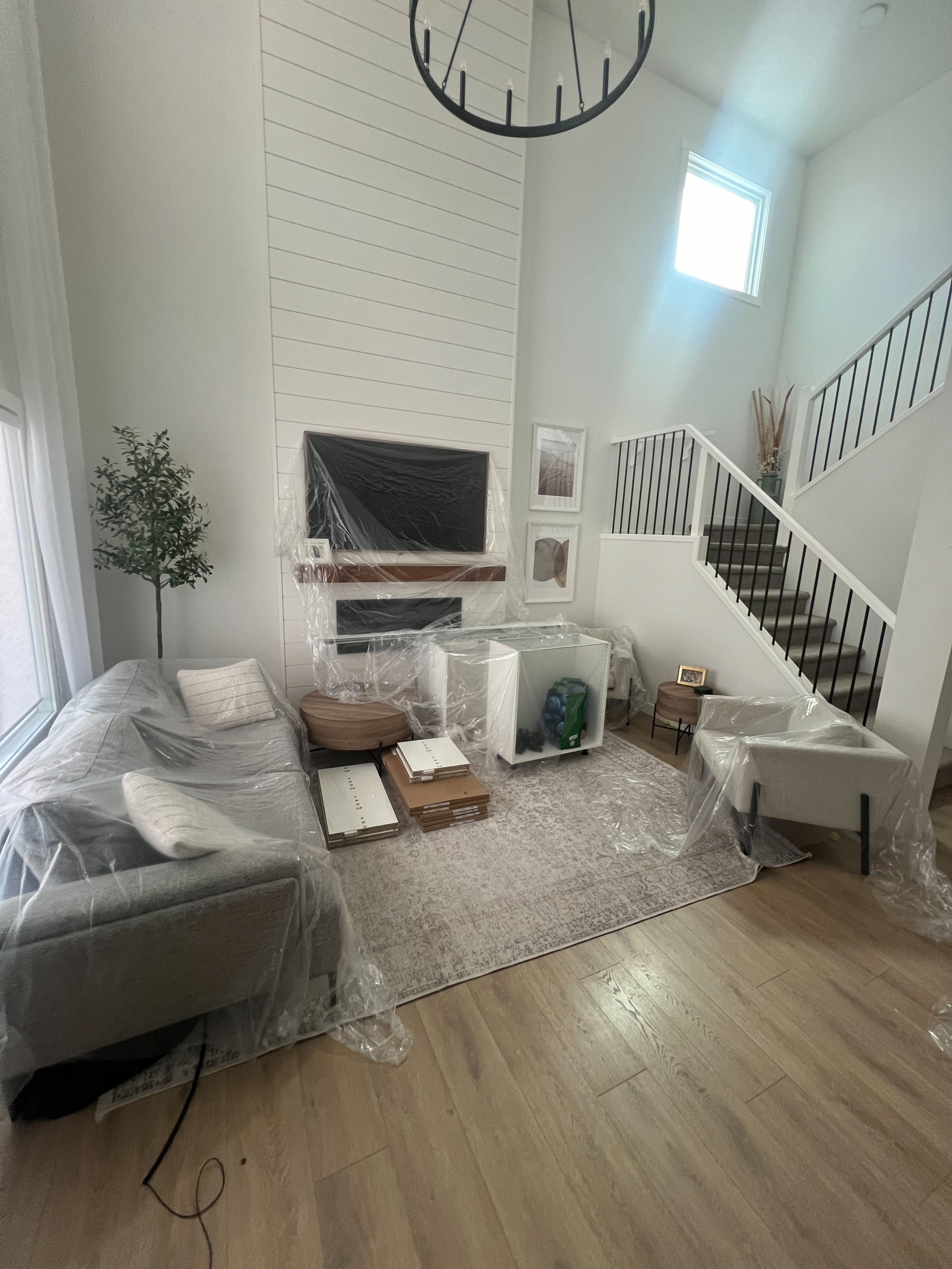 Living room with furniture covered in plastic, featuring a large shiplap wall, a chandelier, staircase, and natural light from a window.
