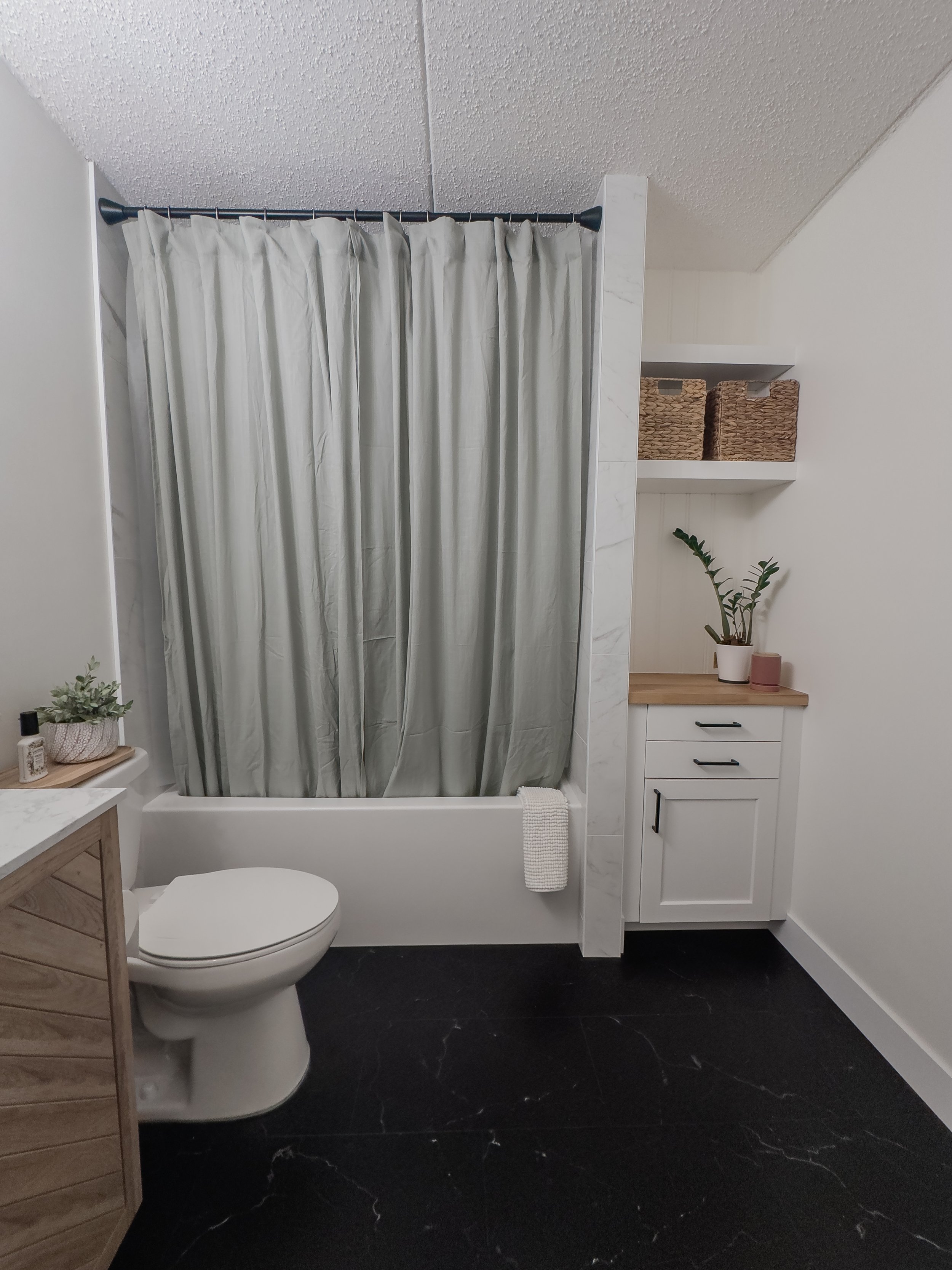 Modern bathroom with a white bathtub and green shower curtain, black floor tiles, white cabinets with potted plants on the counter, and woven baskets on a shelf.