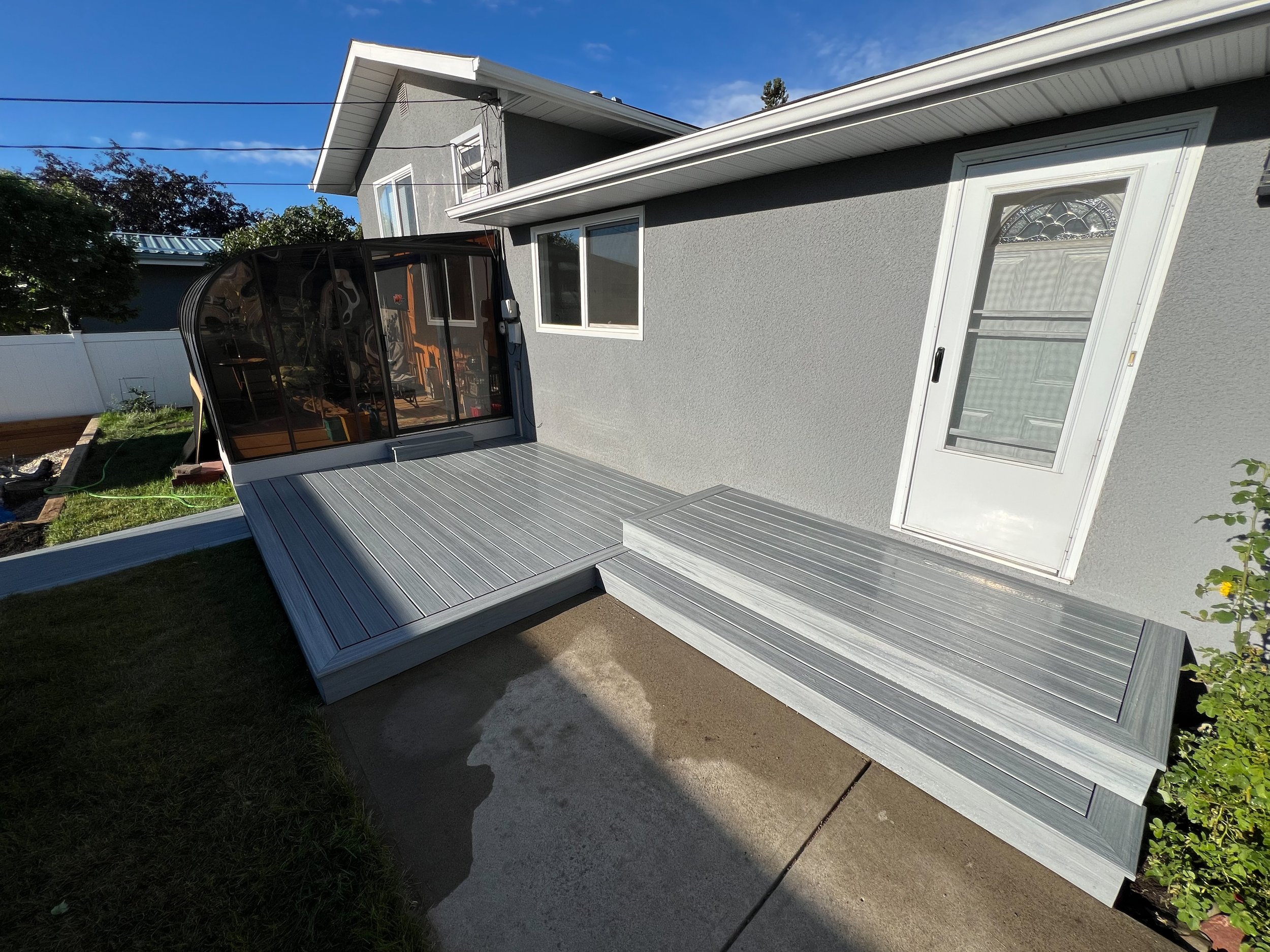 Backyard view of a house with a wooden deck and white door, enclosed patio, grass lawn, and plants.