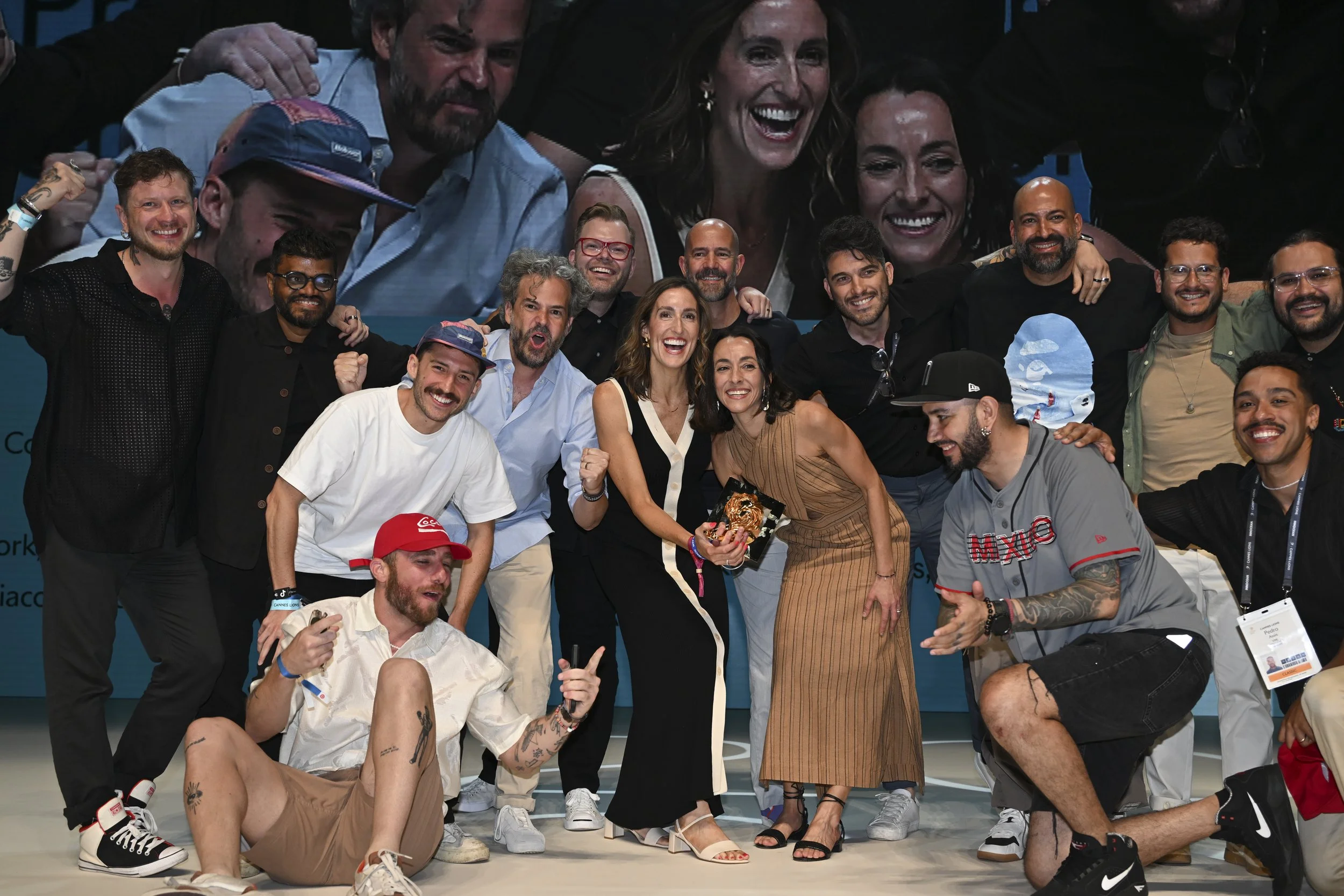 Group of people celebrating on stage at an event, some holding trophies, smiling, and posing for a photo.